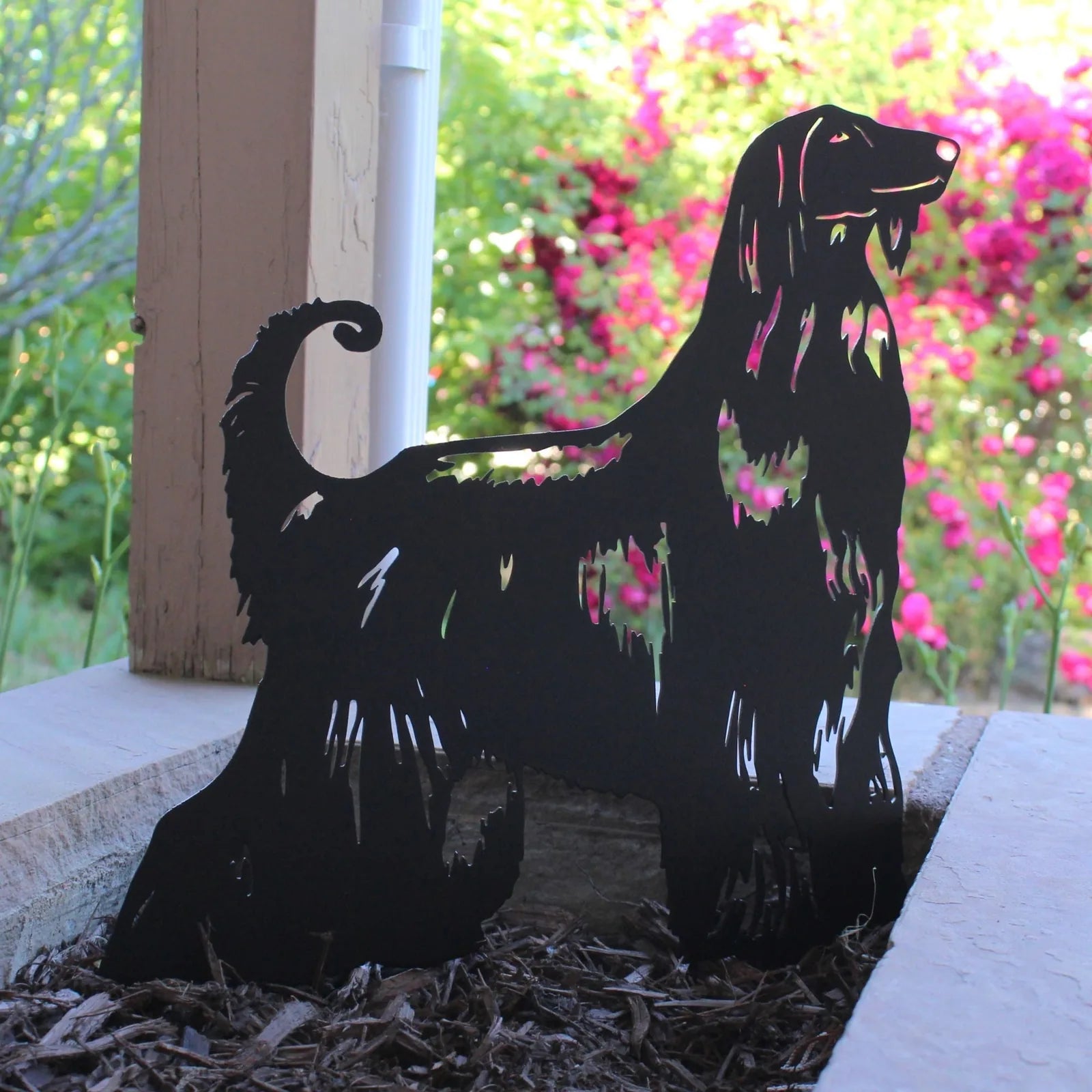 Black Afghan Hound metal silhouette on mulch, surrounded by lush greenery and colorful flowers in the background.