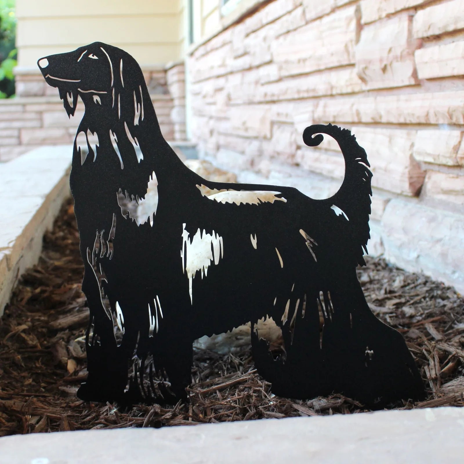 Black metal silhouette of an Afghan Hound, showcasing detailed cutouts, set in wood mulch near a stone wall.