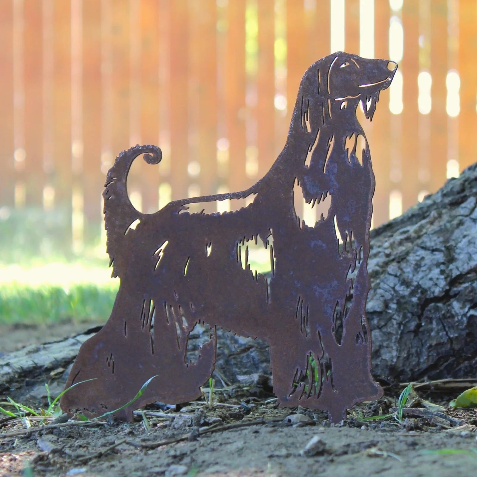 Afghan Hound metal silhouette in weathered Corten steel, standing amidst grass with a wooden fence backdrop.