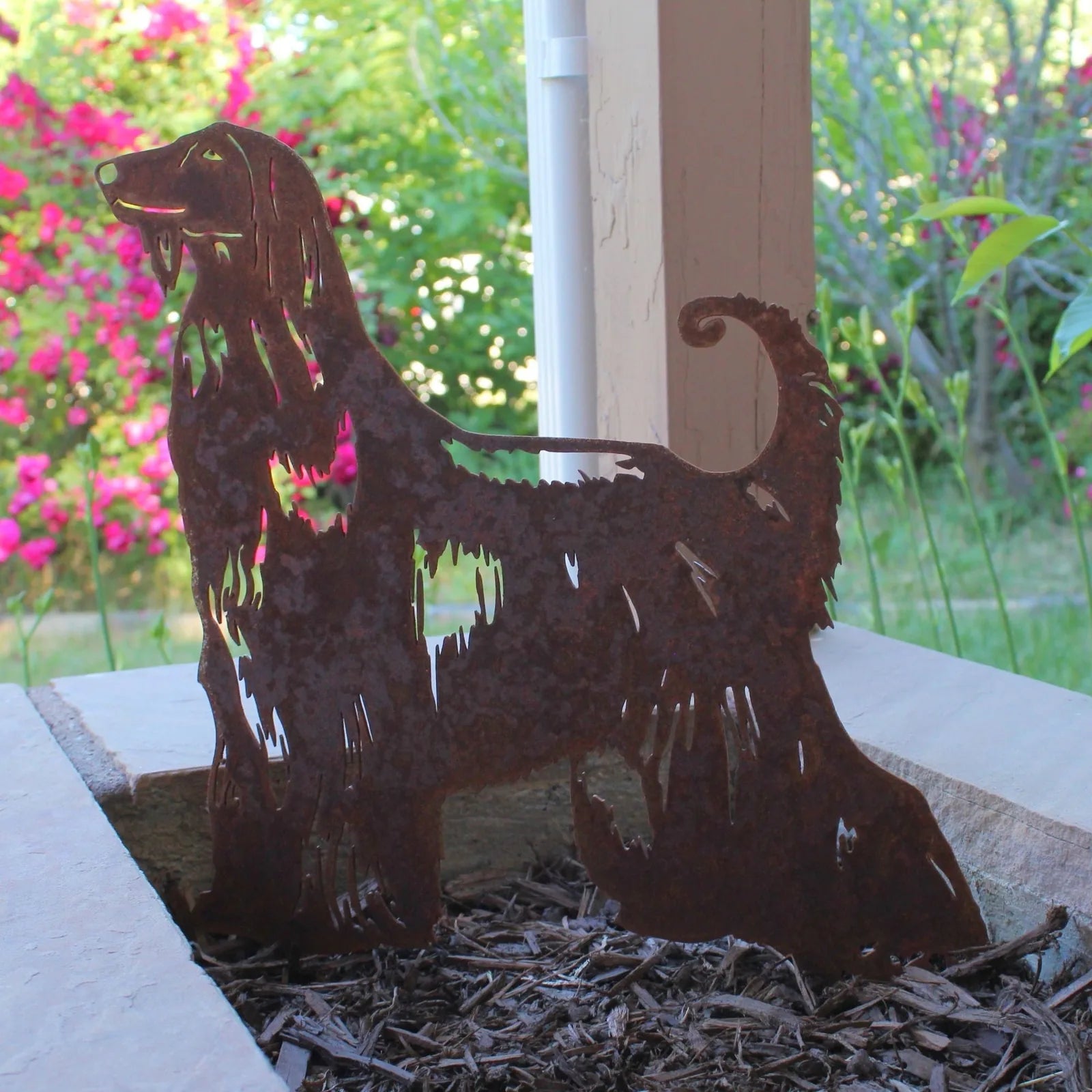 Rusty Corten steel Afghan Hound silhouette standing among mulch with colorful flowers in the background.