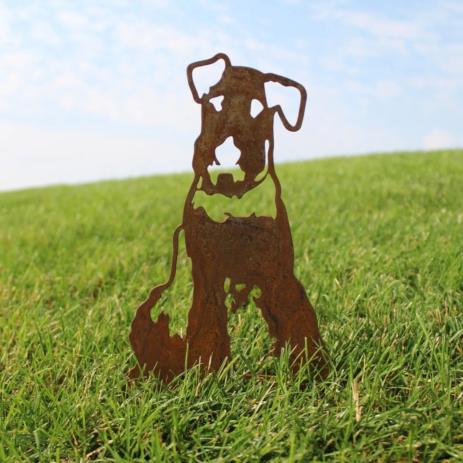 Airedale Terrier silhouette in rusted Corten steel, standing in grass under a bright blue sky.