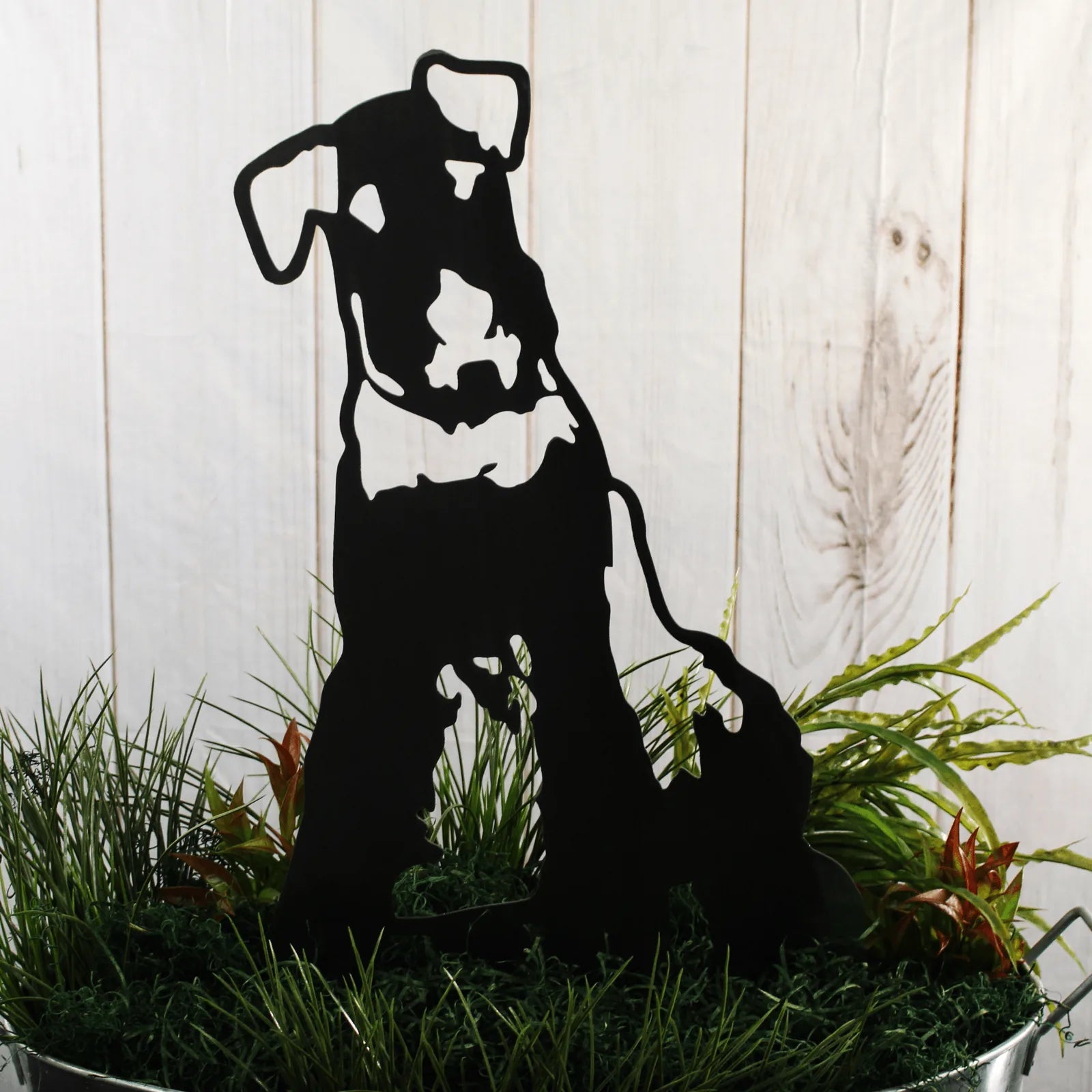 Black metal silhouette of an Airedale Terrier placed among green grass and foliage in an outdoor setting.