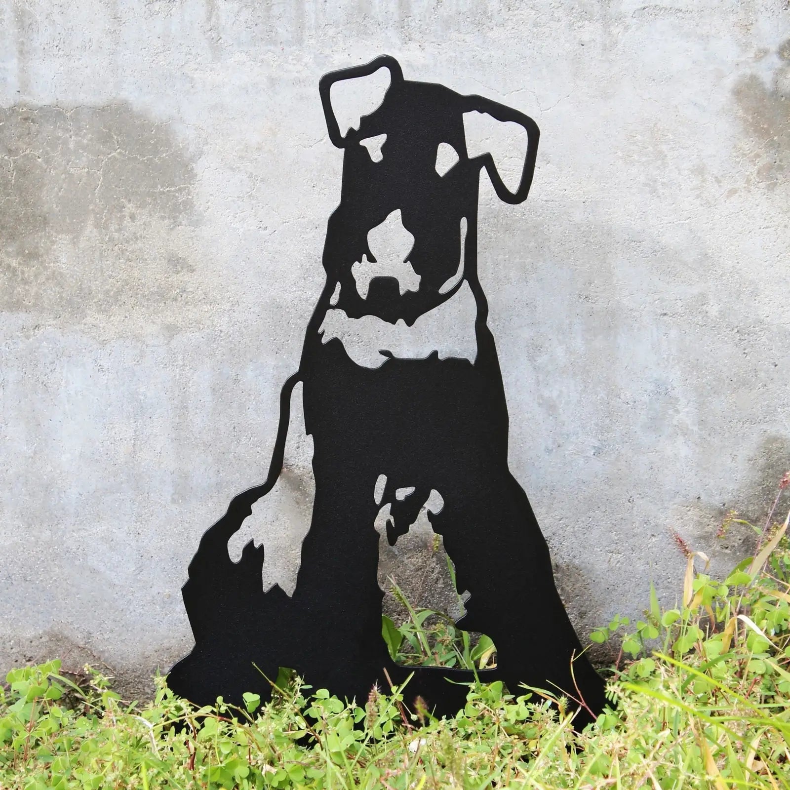 Black Airedale Terrier metal silhouette stands among green foliage against a concrete background, showcasing sleek design.