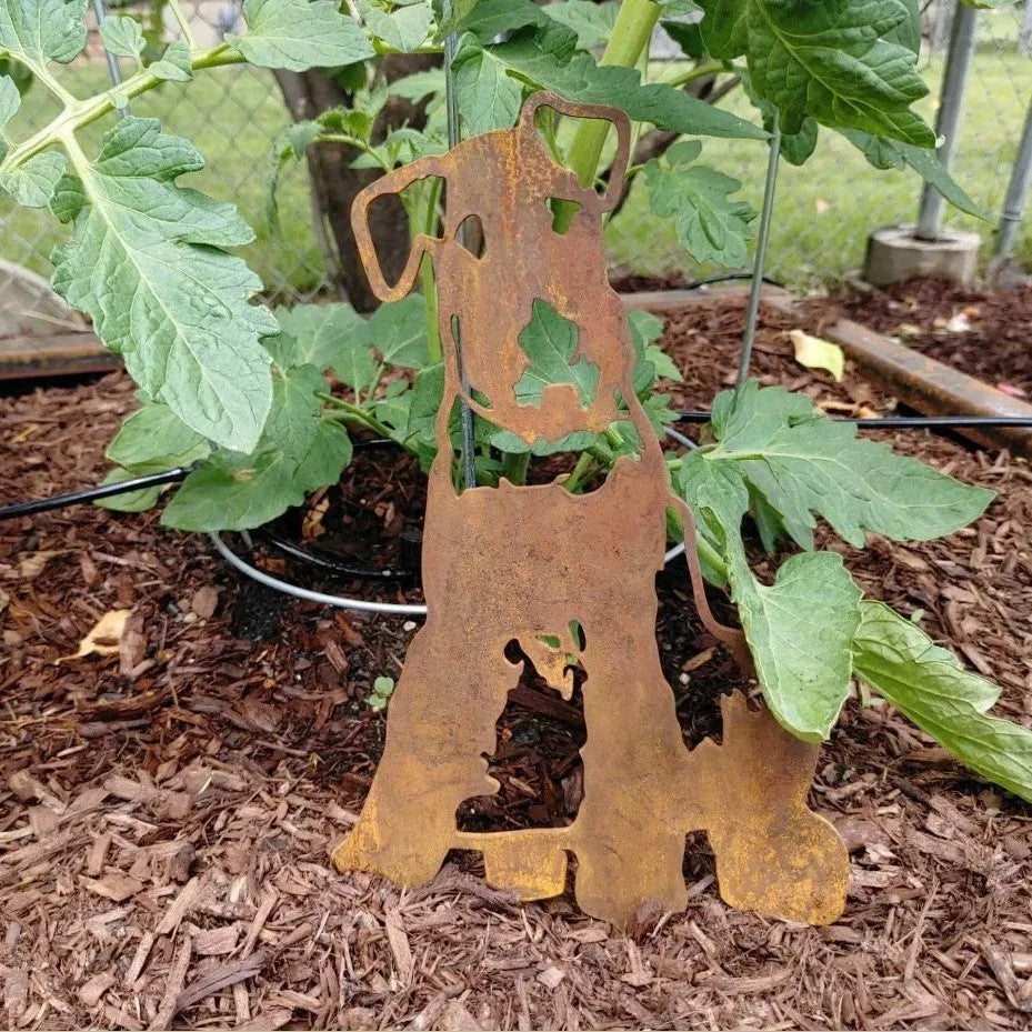 Rusty Airedale Terrier silhouette metal decor, partially hidden by tomato plant leaves, set in mulch.