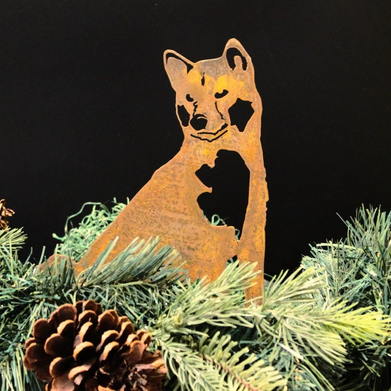 Rusty Akita metal silhouette art piece surrounded by green foliage and pinecones, ideal for outdoor decor.
