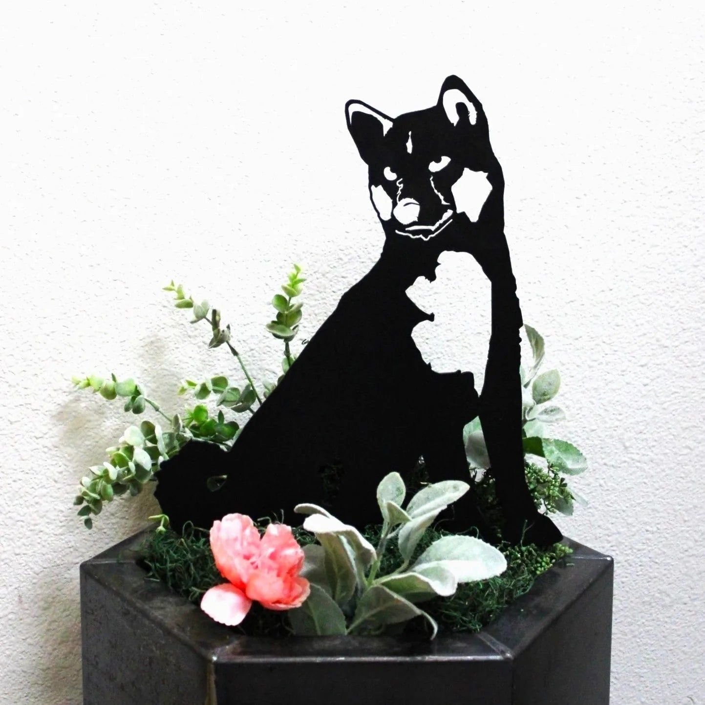 Akita metal silhouette in matte black, nestled among greenery and pink flowers in a planter.