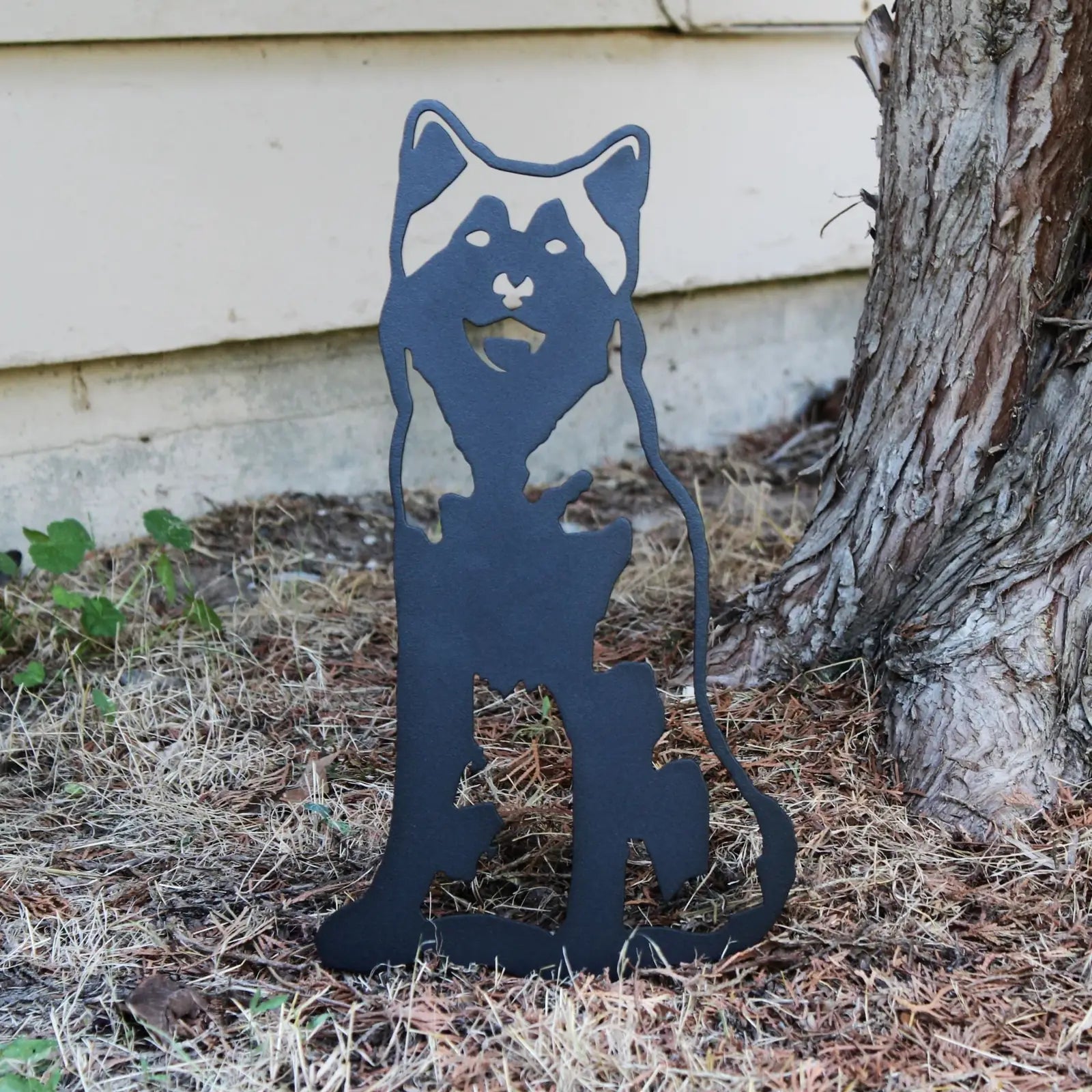 Black metal silhouette of an Alaskan Malamute, standing outdoors among dry grass and tree bark.