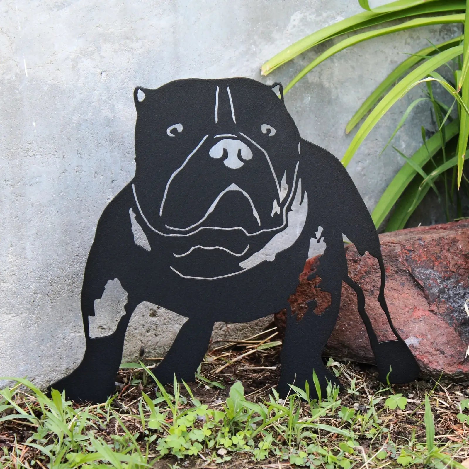 American Bully silhouette in matte black metal, placed outdoors with greenery and rocks, showcasing its sturdy design.