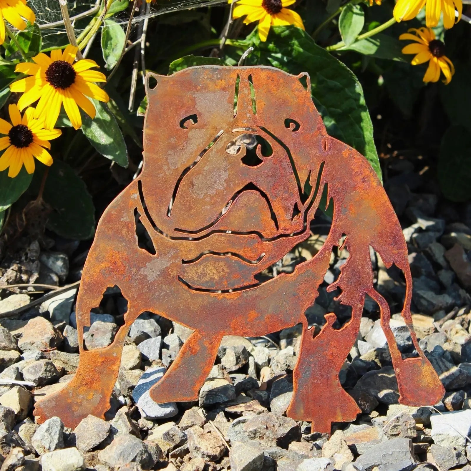 Rusty Corten steel silhouette of an American Bully among yellow flowers and gravel, showcasing unique patina and design.