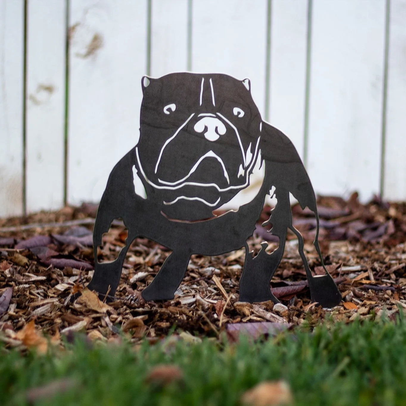 American Bully silhouette in matte black metal, displayed on mulch in a garden setting.