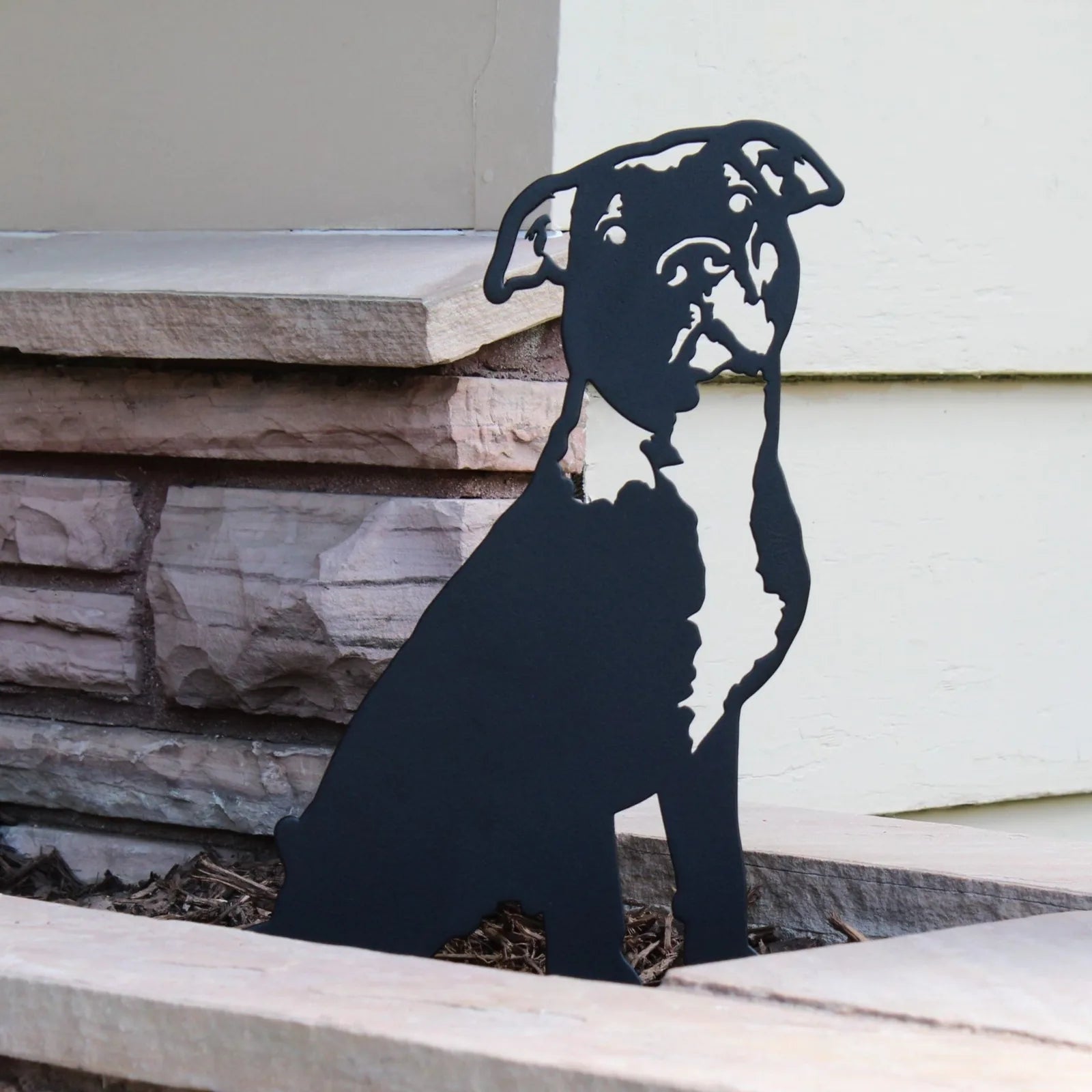 Black metal silhouette of an American Pit Bull Terrier, displayed in landscaping next to a stone wall.