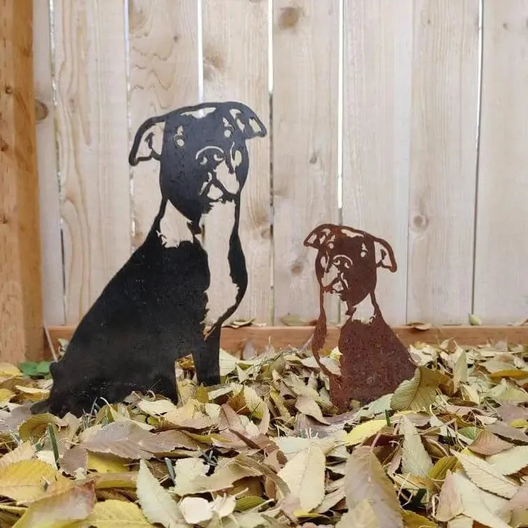 Black and Corten steel silhouettes of American Pit Bull Terriers among fallen leaves, set against a wooden fence.