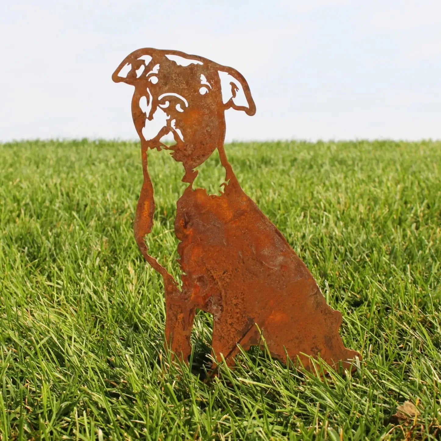 Rusty Corten steel silhouette of an American Pit Bull Terrier, set in green grass against a clear sky.