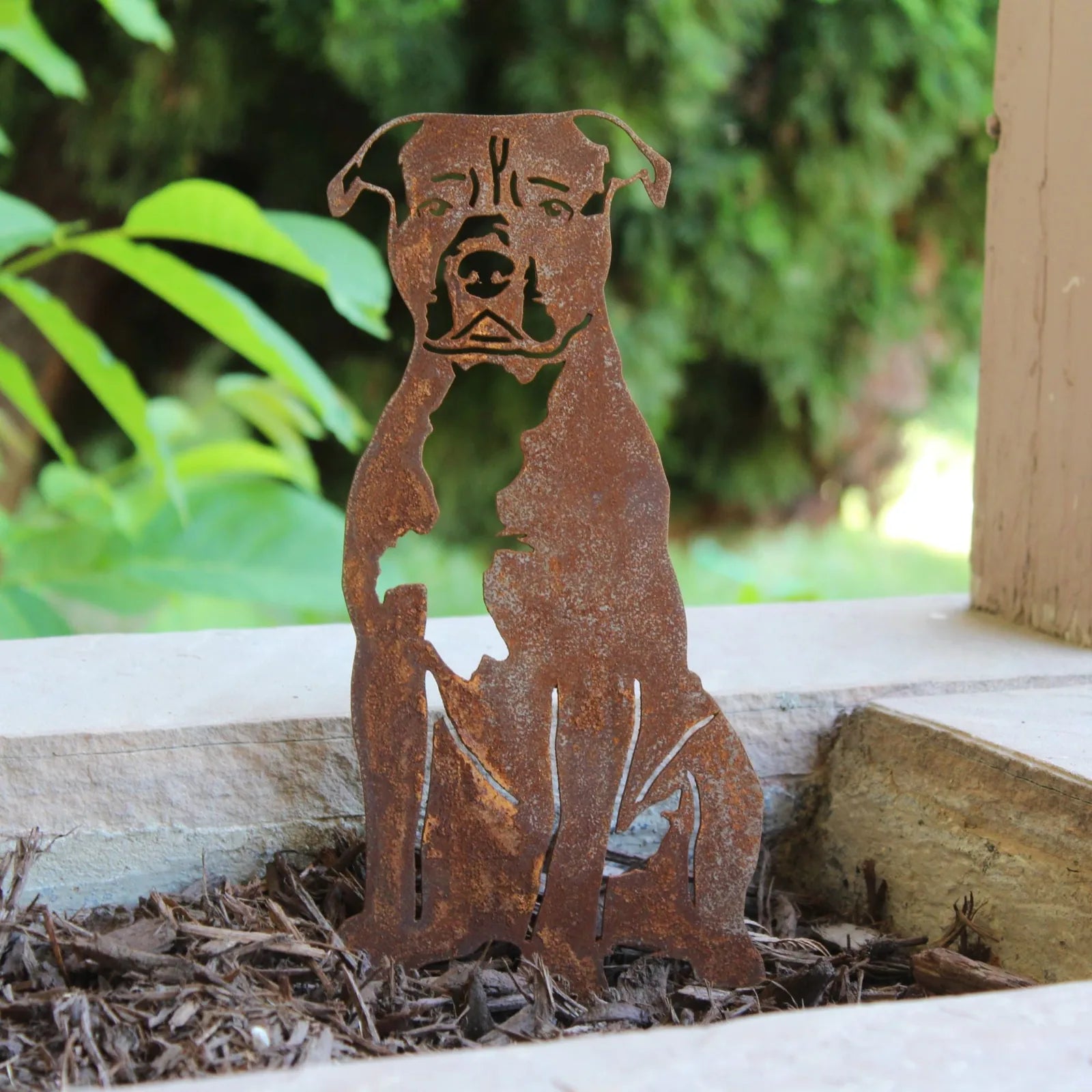 Handcrafted rusted metal silhouette of an American Staffordshire Terrier, displayed in mulch against a garden backdrop.