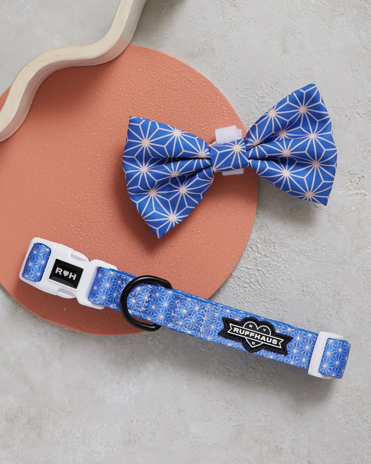 Blue dog collar with geometric pattern and detachable bowtie, displayed on a round orange surface.