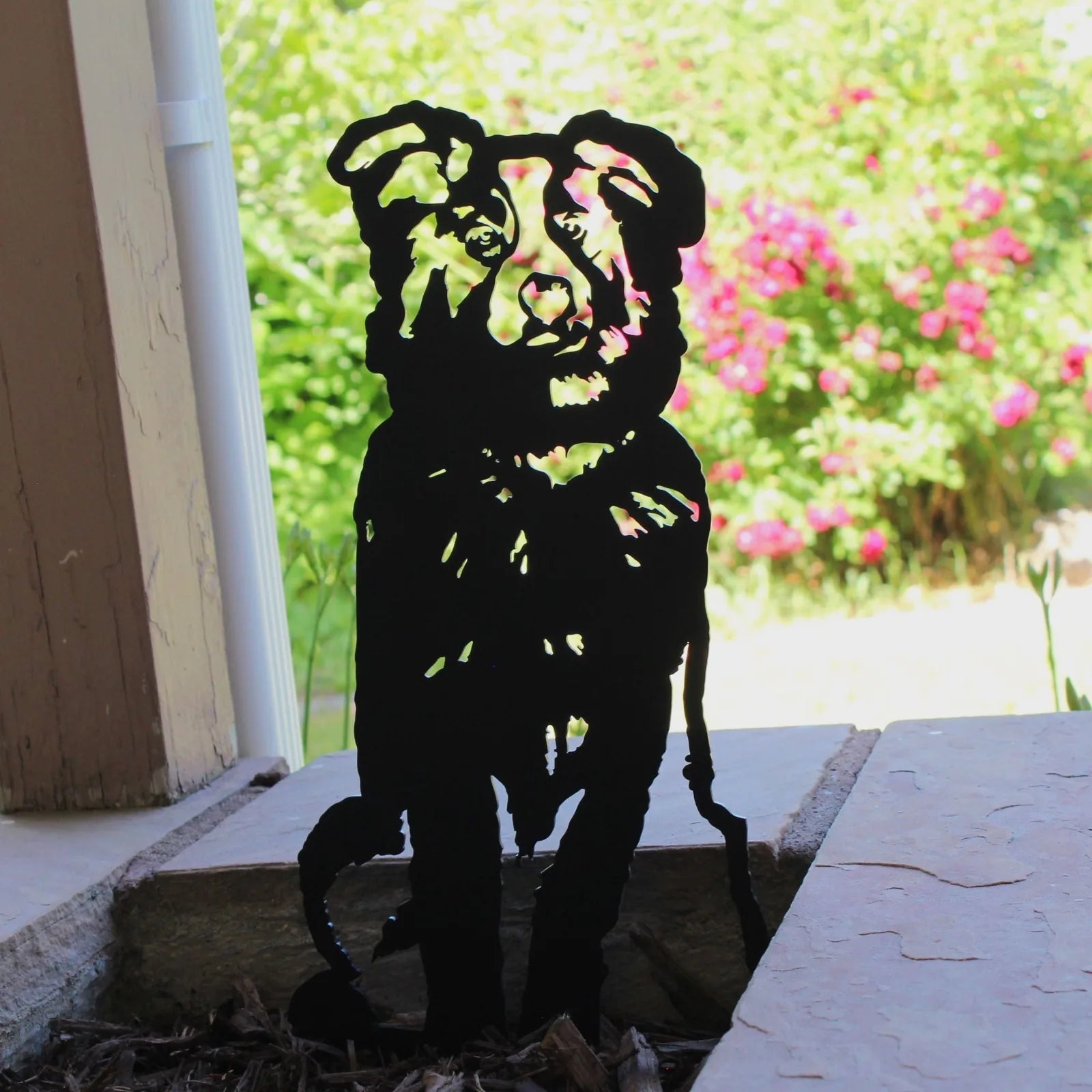 Black Australian Shepherd metal silhouette standing on stone, surrounded by lush greenery and colorful flowers.