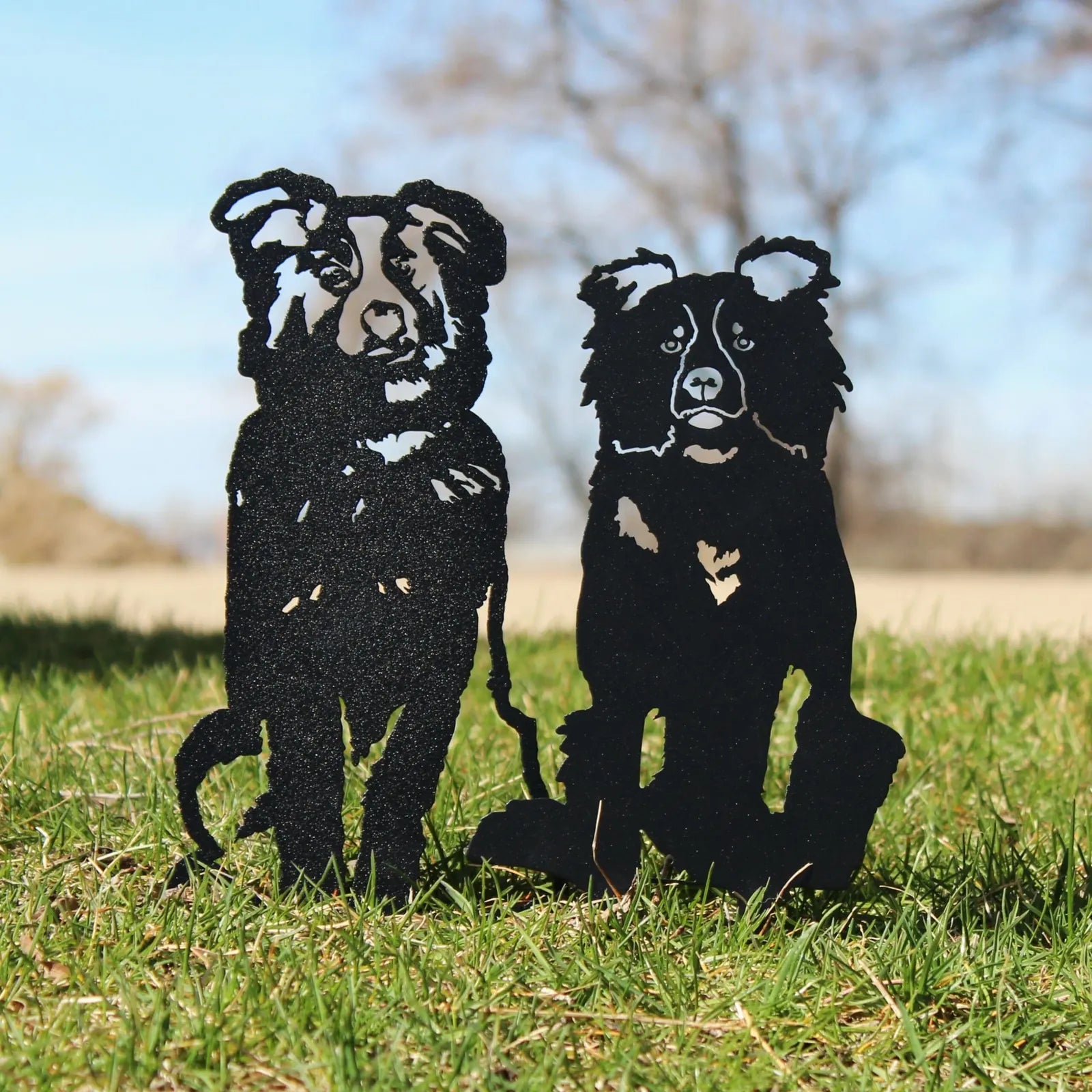 Black metal silhouettes of two Australian Shepherds sitting on grass with a clear sky in the background.