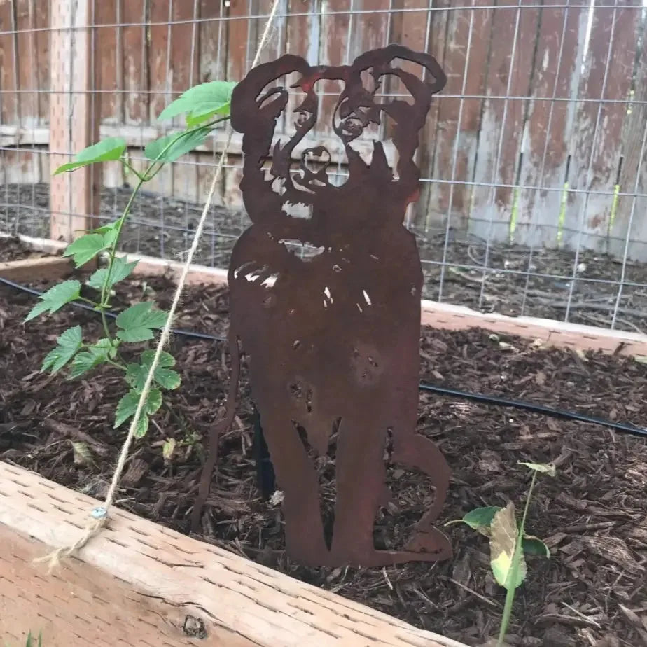 Rust-colored Australian Shepherd metal silhouette standing in a garden bed surrounded by mulch and plants.