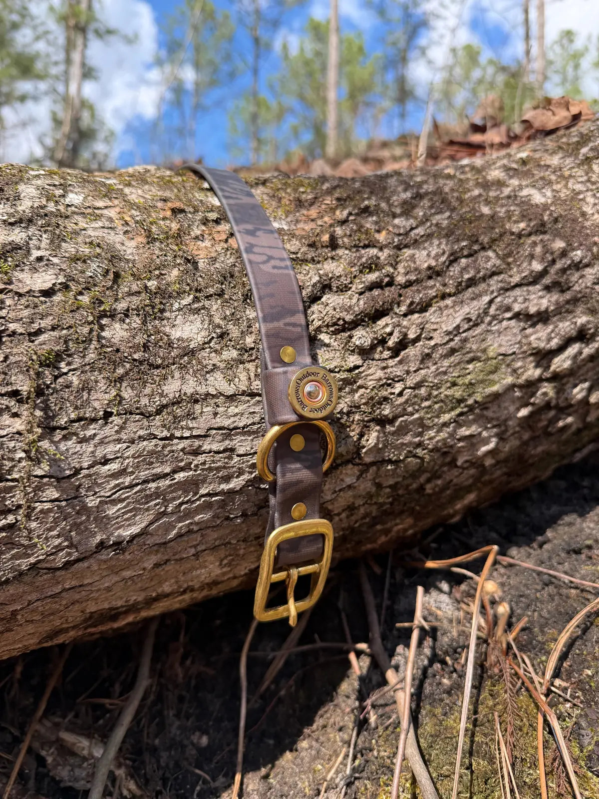 Brown camo Barrows dog collar resting on a fallen log, featuring brass hardware and Biothane material for durability.