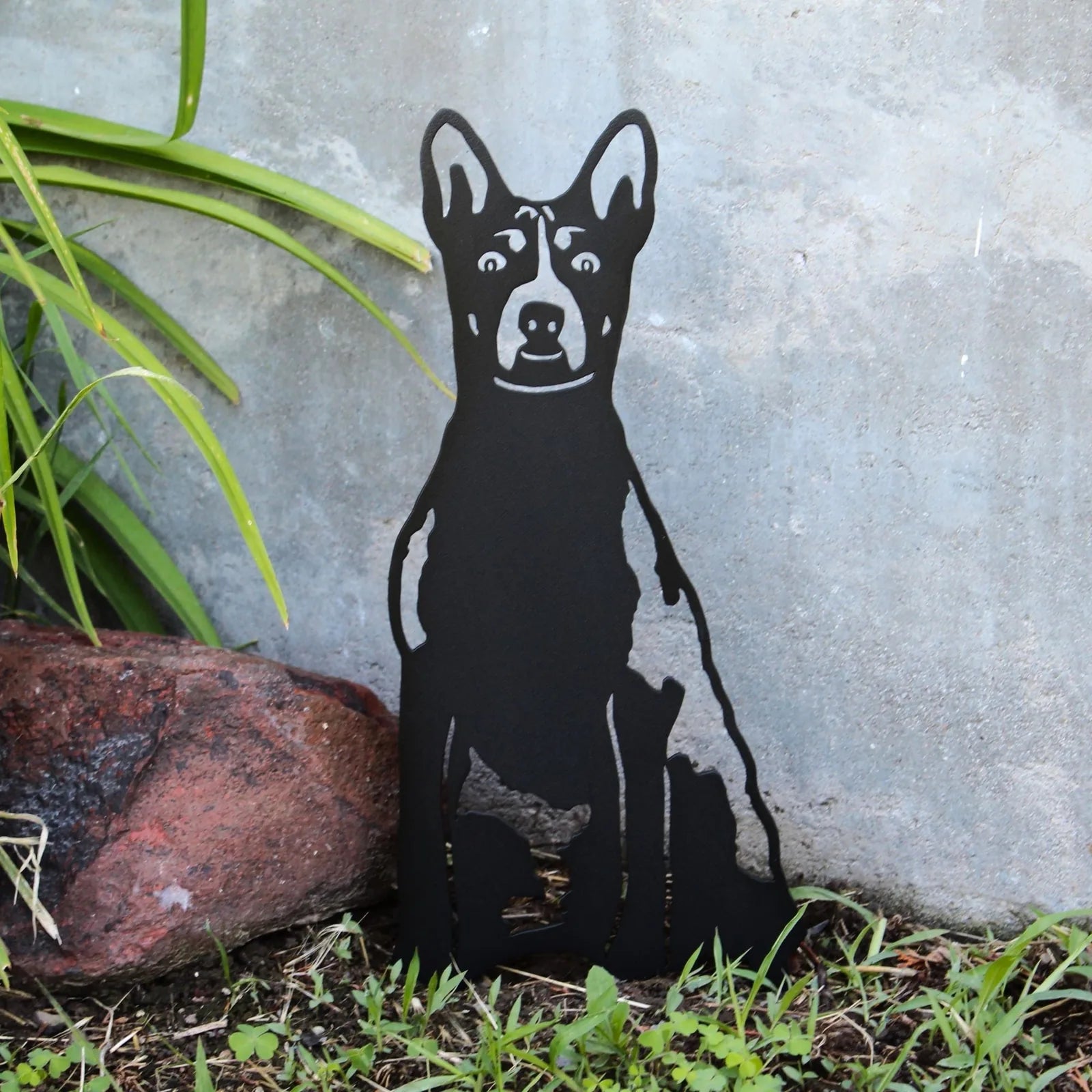 Black metal silhouette of a Basenji dog sitting beside gray stone and green grass, ideal for outdoor decor.