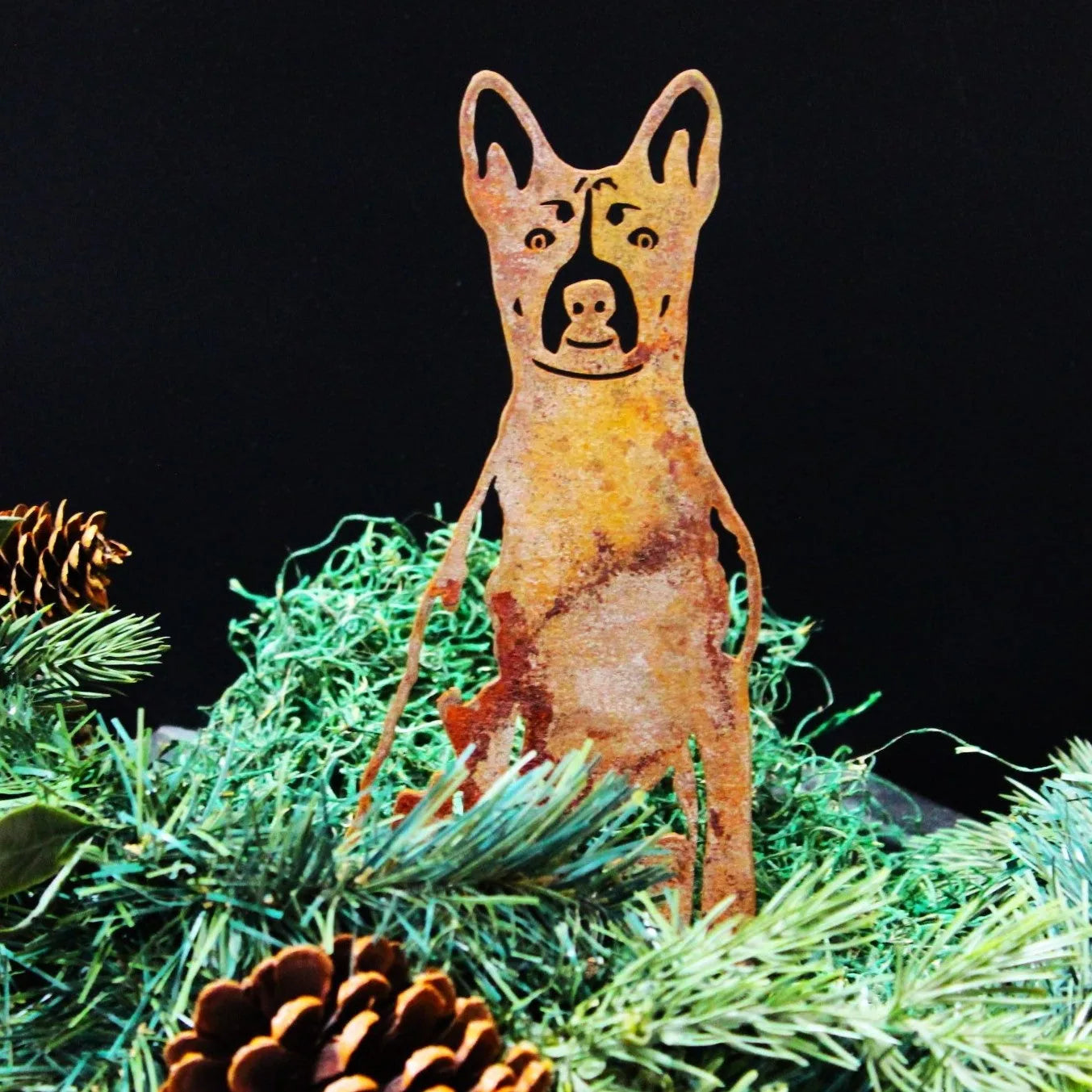 Rusty Basenji metal silhouette surrounded by green foliage and pinecones, showcasing its weathered Corten steel design.