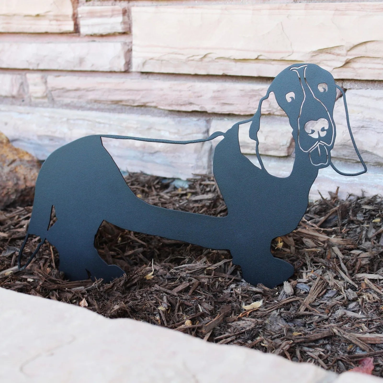 Black metal silhouette of a Basset Hound, detailed design resting in garden mulch, blending with outdoor decor.