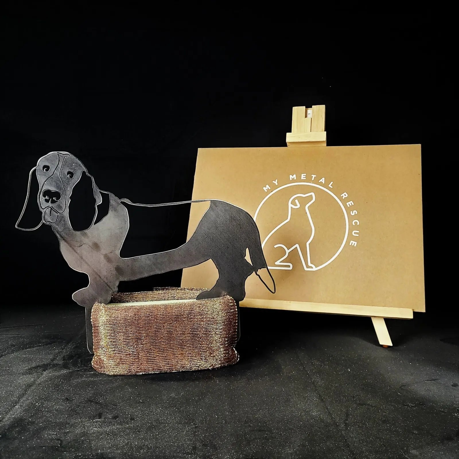 Basset Hound metal silhouette in matte black, displayed on a textured base with a branded backdrop.