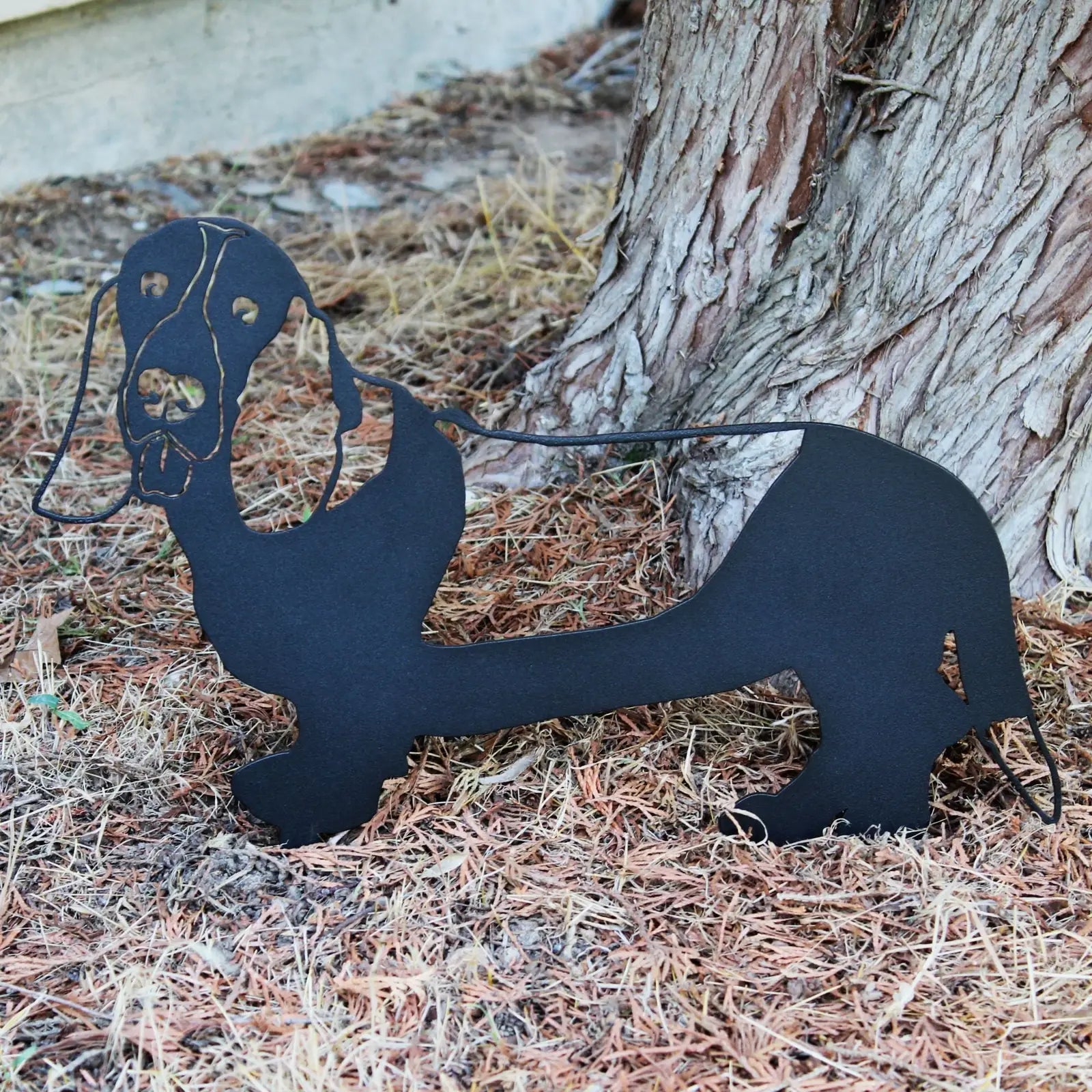 Black Basset Hound metal silhouette sculpture resting on pine needles near a tree, ideal for garden decor.