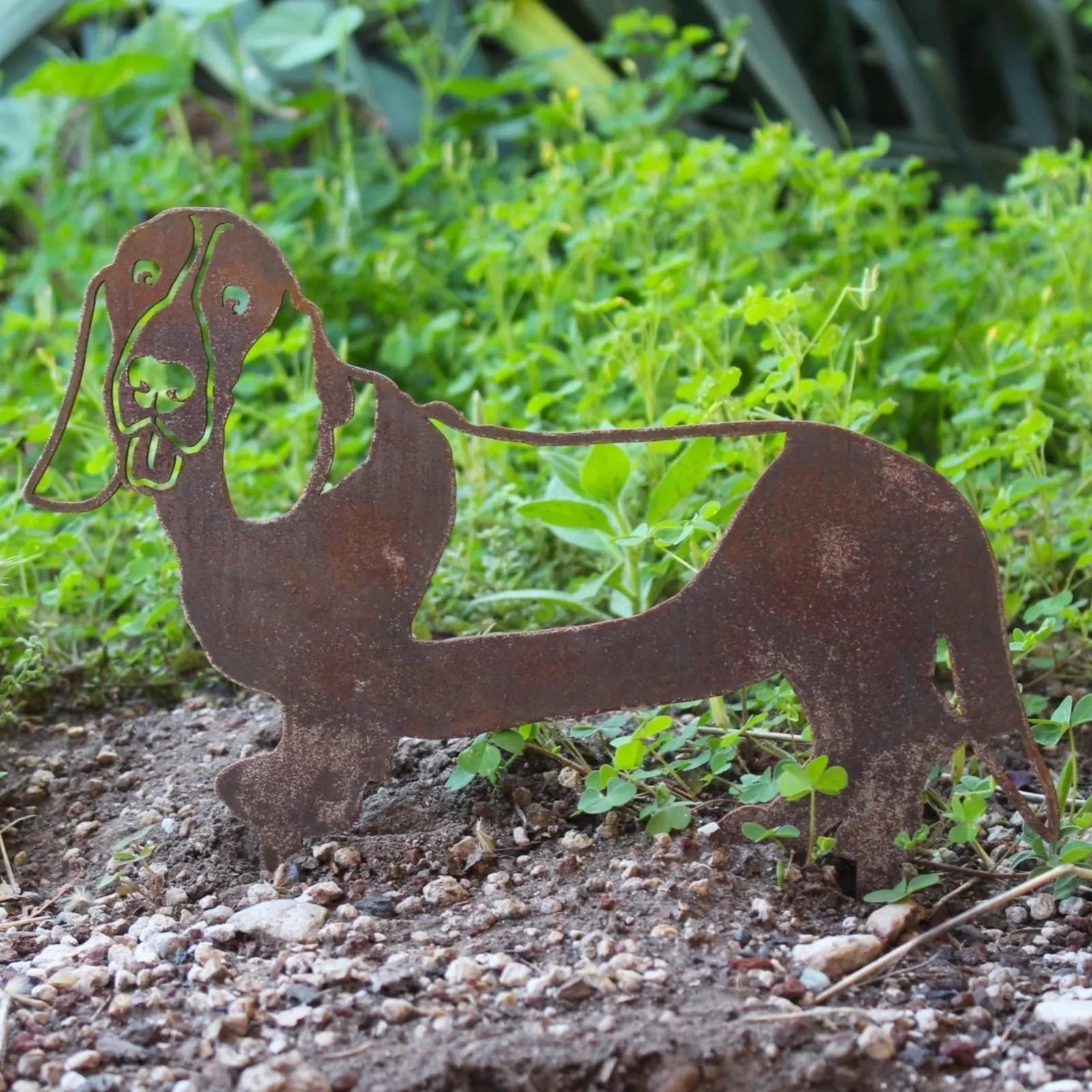 Rusty Basset Hound silhouette made of Corten steel, set against green garden foliage, enhancing outdoor decor.