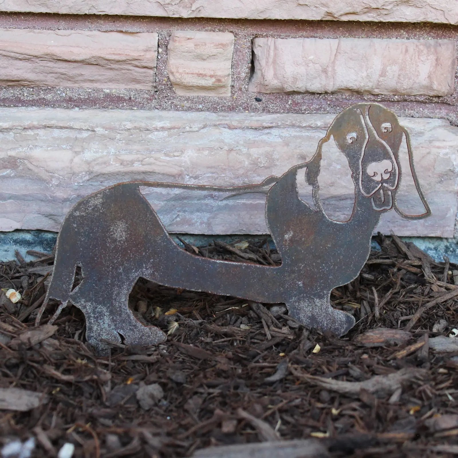 Basset Hound metal silhouette in rusted Corten steel, displayed against mulch and a stone wall background.