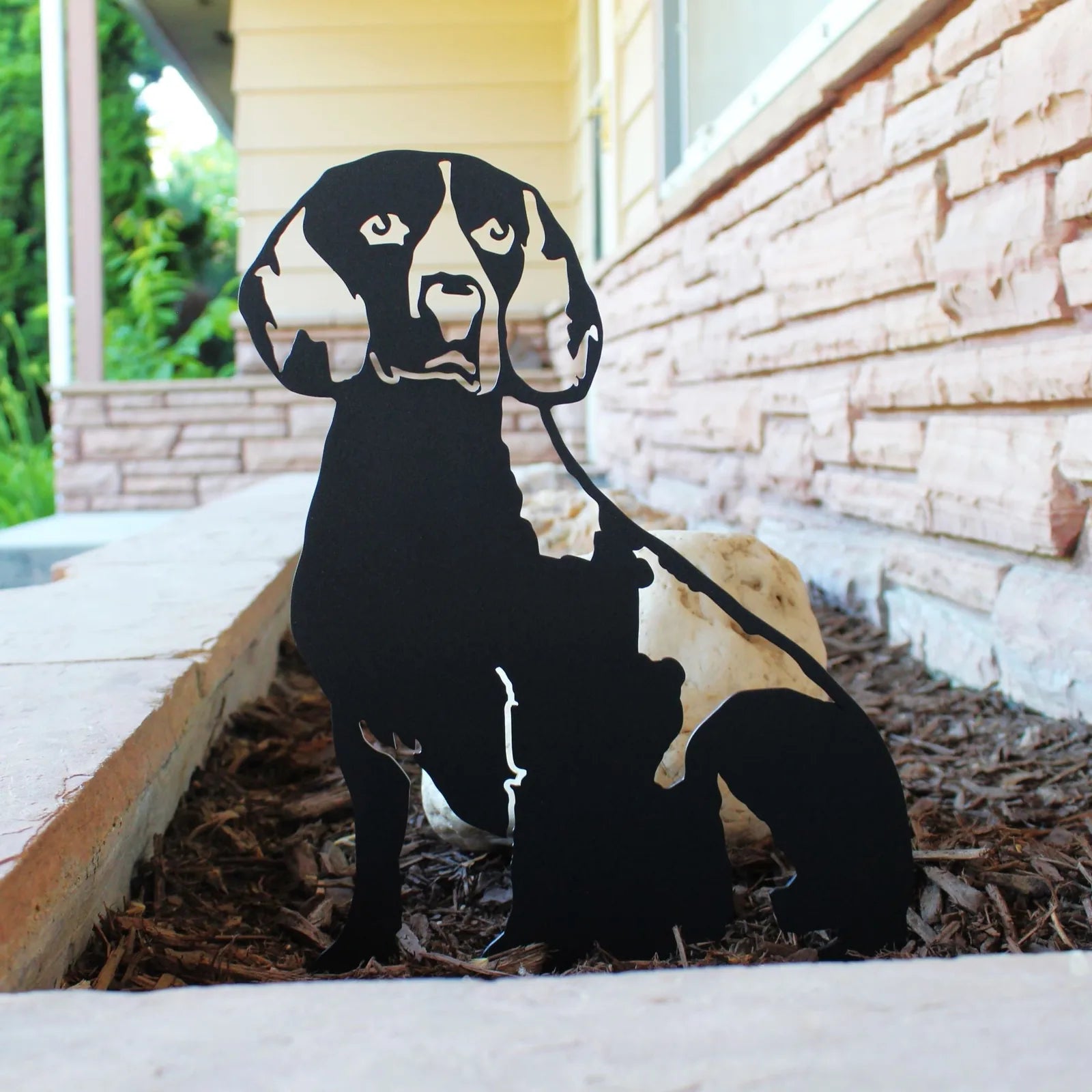 Beagle metal silhouette in matte black, displayed in mulch against a stone backdrop, adding rustic dog decor.