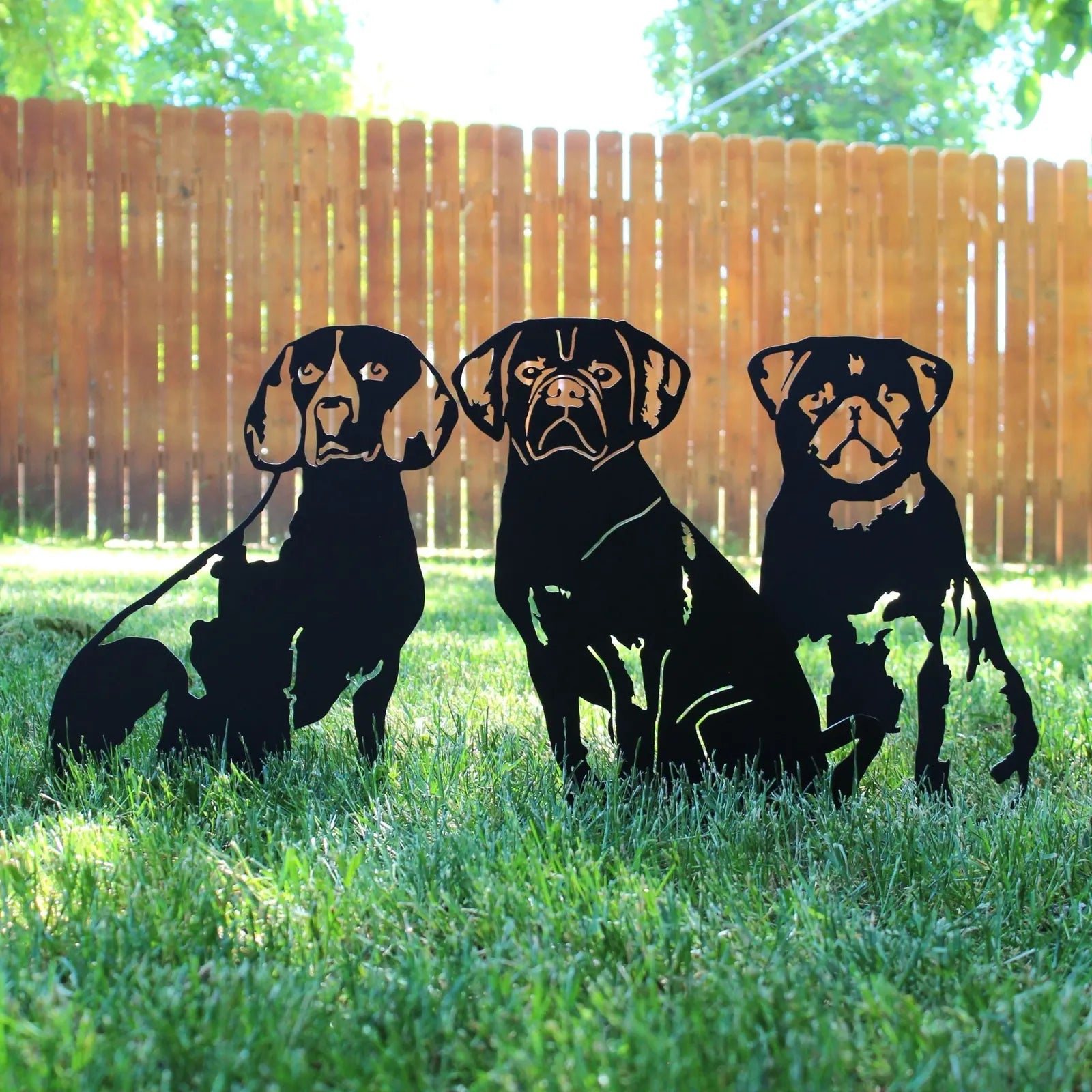 Black metal silhouettes of three dogs in a grassy yard, showcasing detailed shapes and expressions, with a wooden fence be...