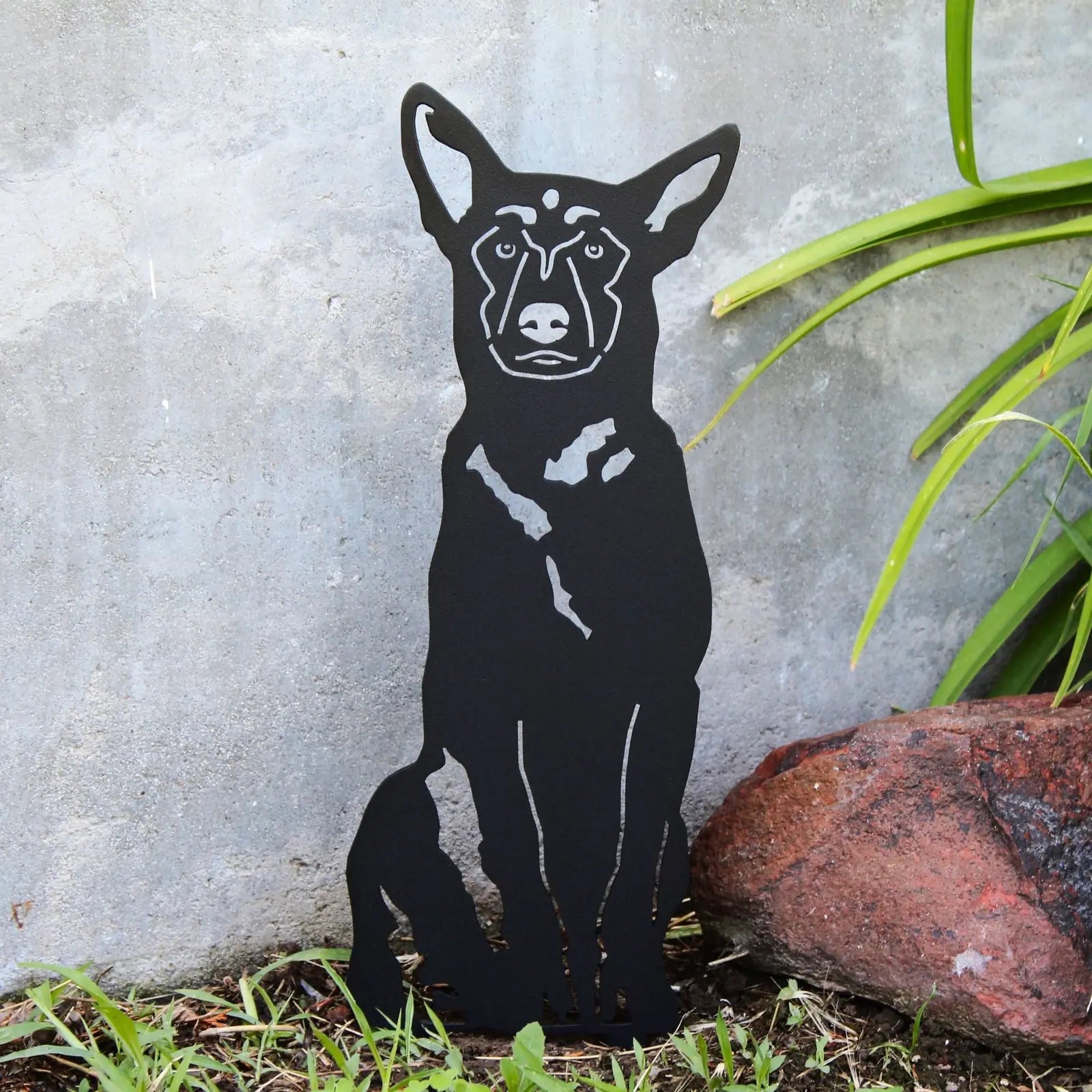 Belgian Malinois metal silhouette in matte black, positioned among greenery and rocks against a concrete background.
