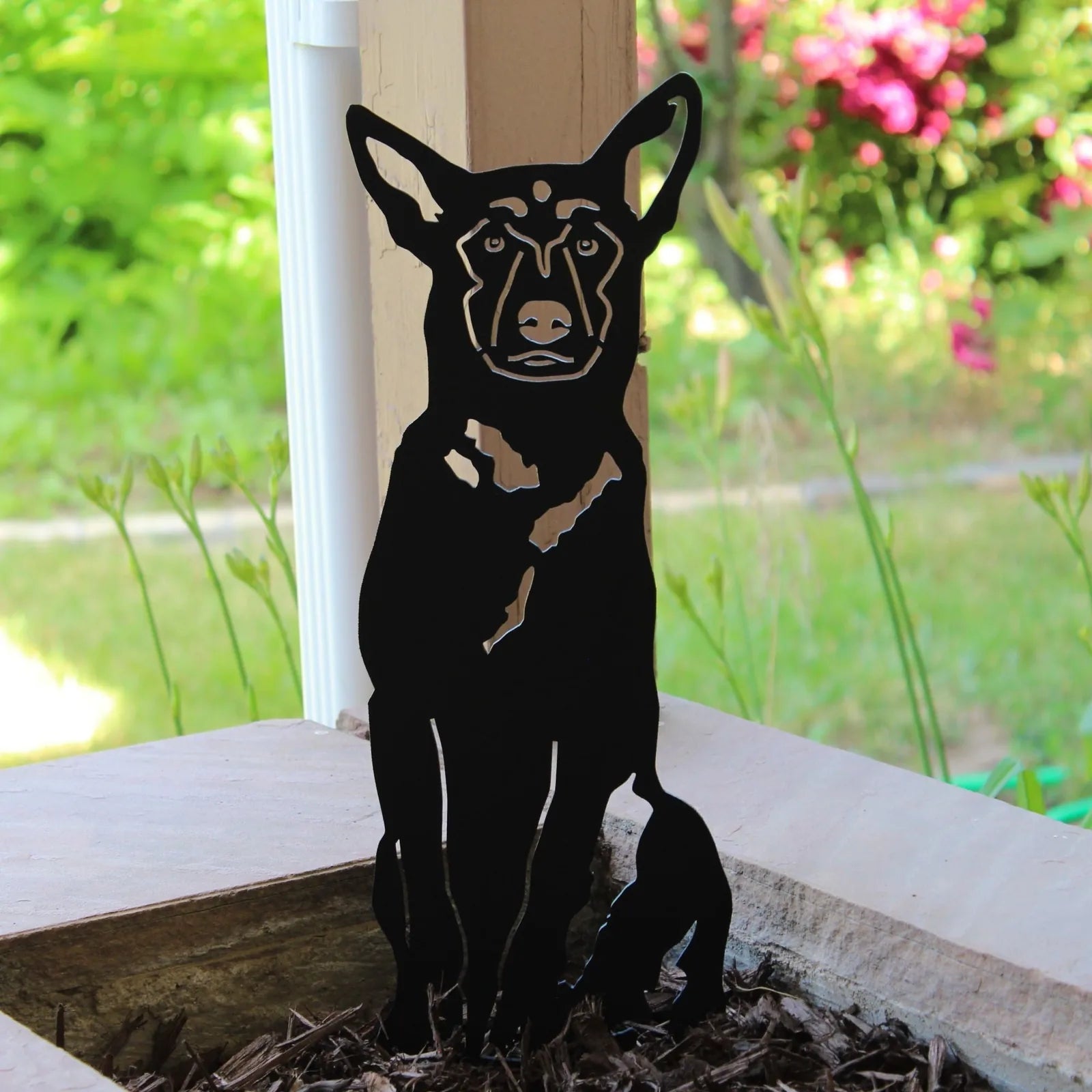 Black metal silhouette of a Belgian Malinois dog, displaying intricate cutouts, placed in a garden setting.
