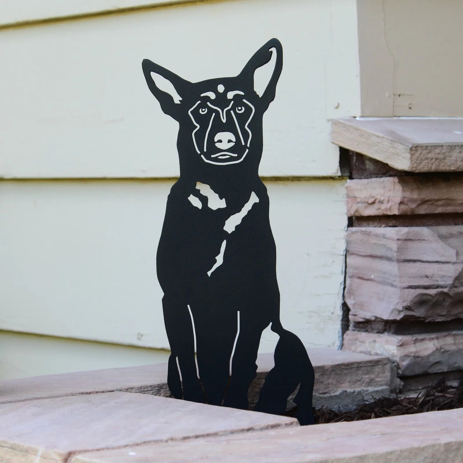 Black Belgian Malinois metal silhouette art piece, standing against a light-colored wall and stone backdrop.