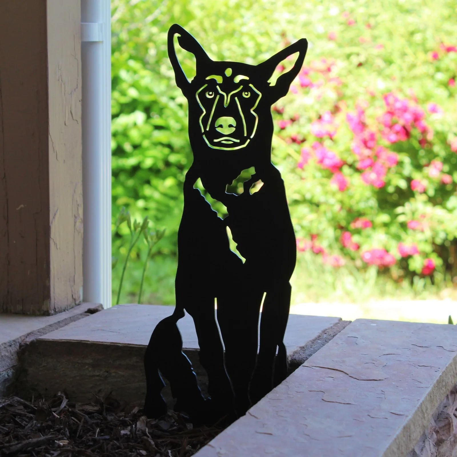 Black metal silhouette of a Belgian Malinois sits on stone, surrounded by green foliage and pink flowers.
