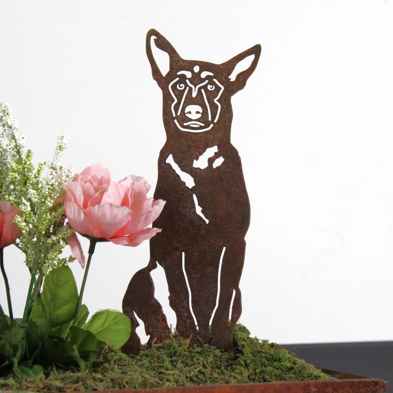 Belgian Malinois metal silhouette in rust finish, set among pink flowers and greenery in a decorative arrangement.
