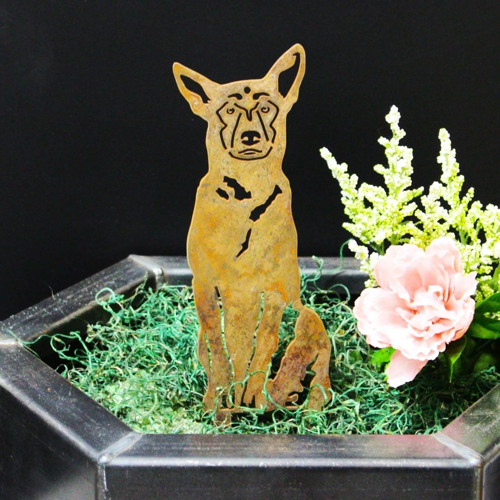 Belgian Malinois metal silhouette in weathered steel, showcasing detailed cutouts, surrounded by greenery and flowers.