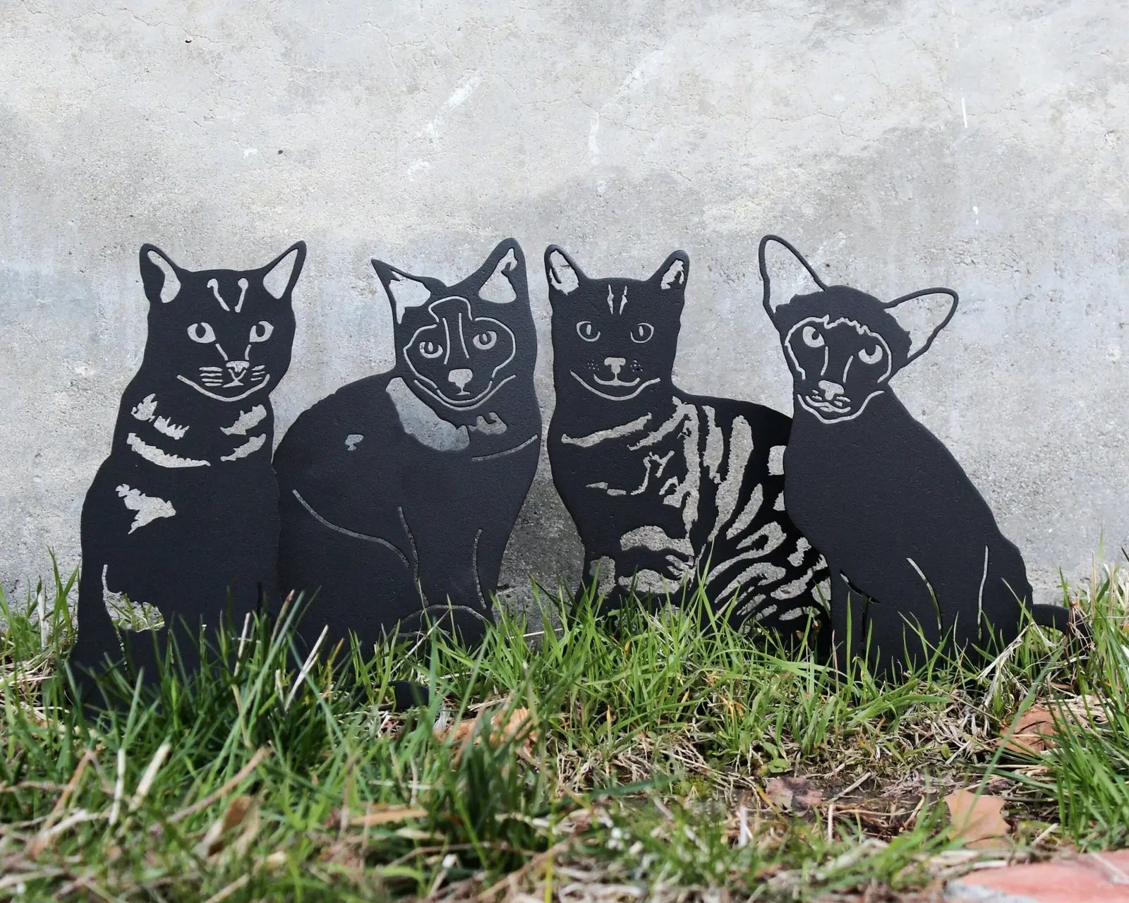 Black metal silhouette art depicting four distinct cat designs, set against a concrete backdrop with grass.