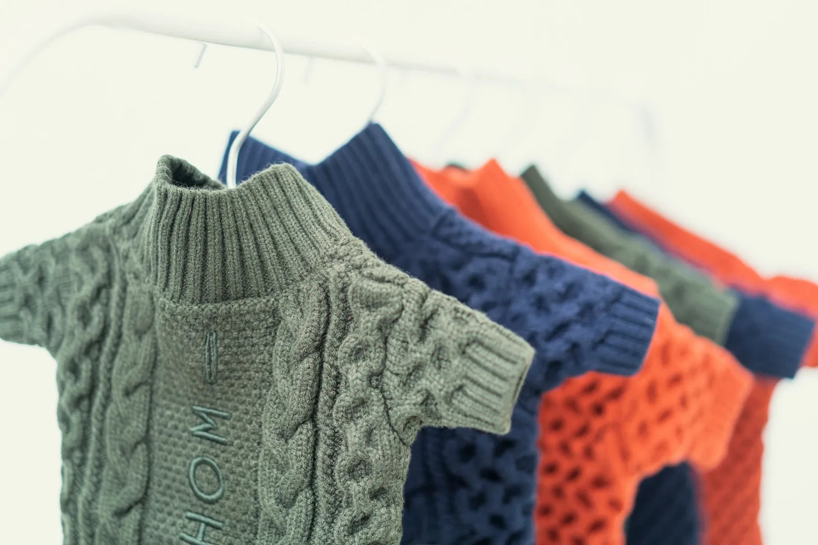 Orange BERT Cardigan Sweater for dogs, featuring a cable knit design and ribbed collar, hanging alongside other colors.