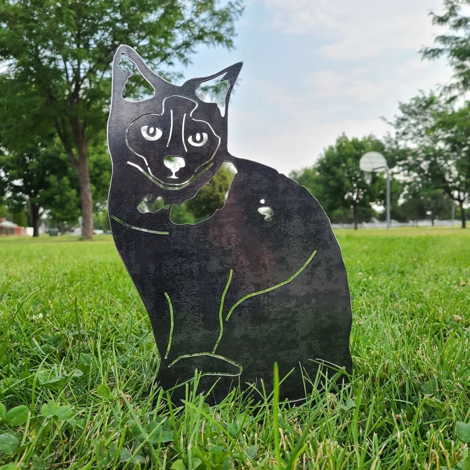 Birman cat silhouette in black metal, sitting among vibrant green grass under a clear sky.