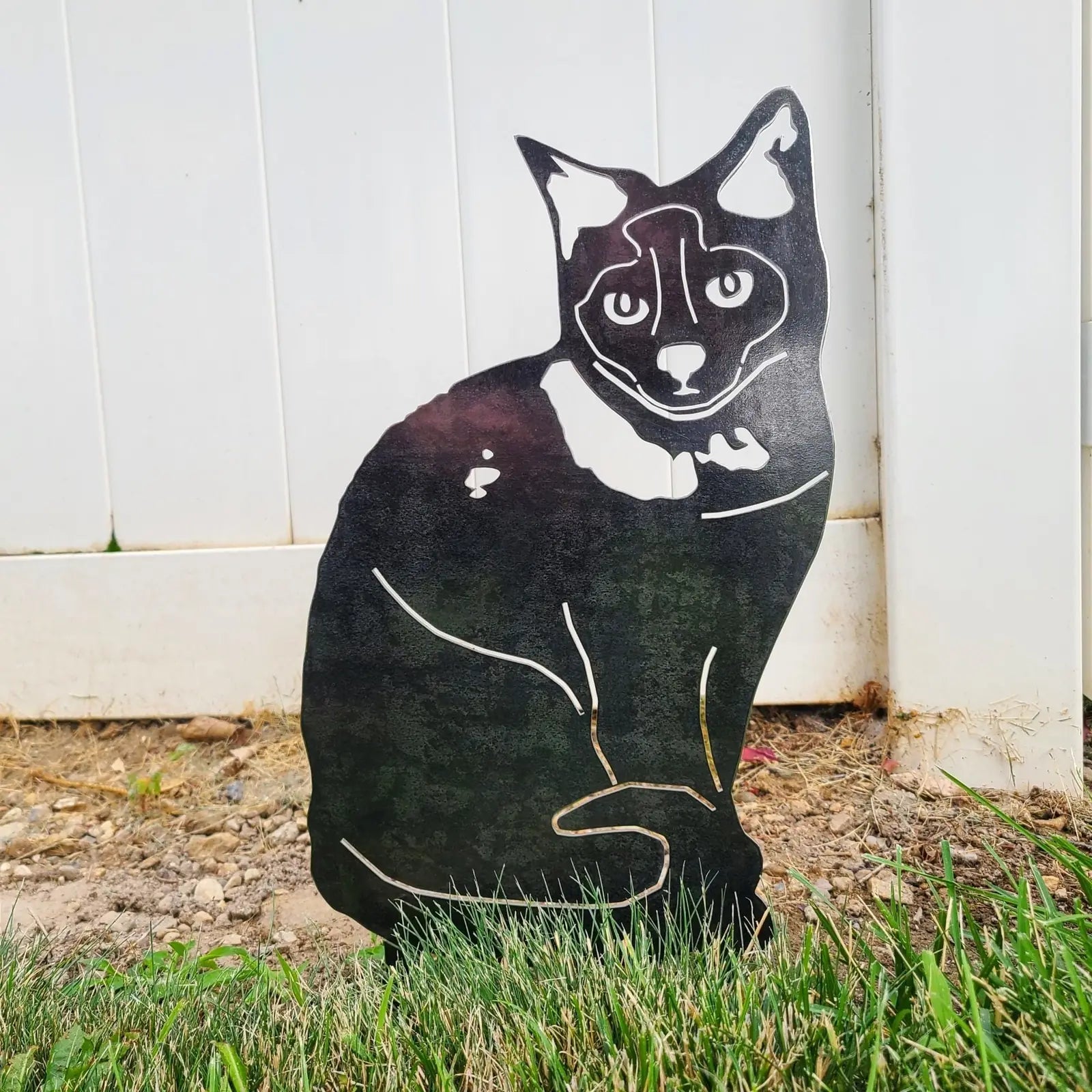 Black metal silhouette of a sitting Birman cat, designed for outdoor display, with grass and a white fence in the background.