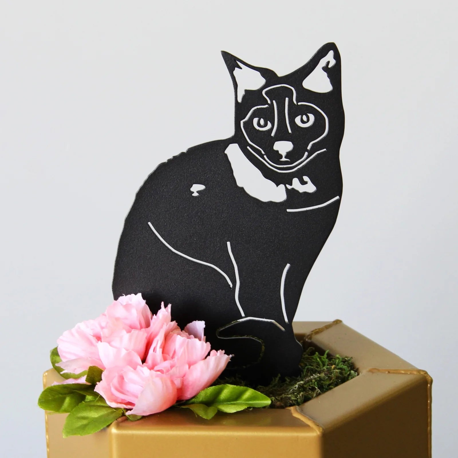 Black metal silhouette of a Birman cat with white accents, placed among pink flowers in a gold planter.