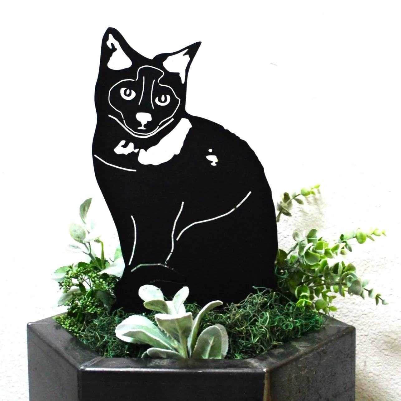 Black metal silhouette of a Birman cat sitting amidst green foliage in a decorative planter.