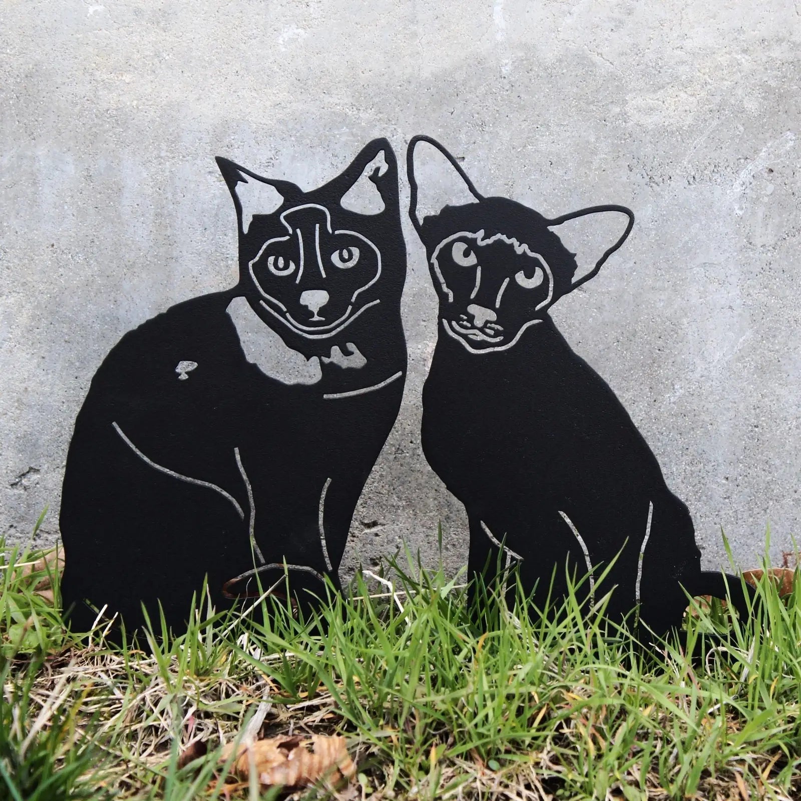 Black metal silhouette of two cats, showcasing detailed features against a stone backdrop, with grass at the base.