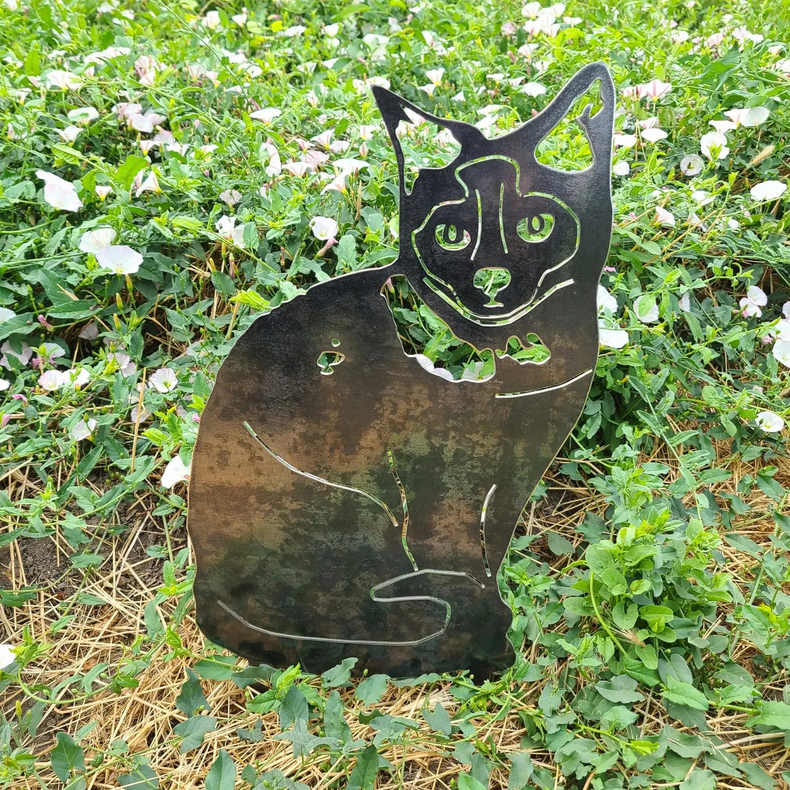Birman metal silhouette art display in weathered Corten steel, surrounded by green foliage and pink flowers.