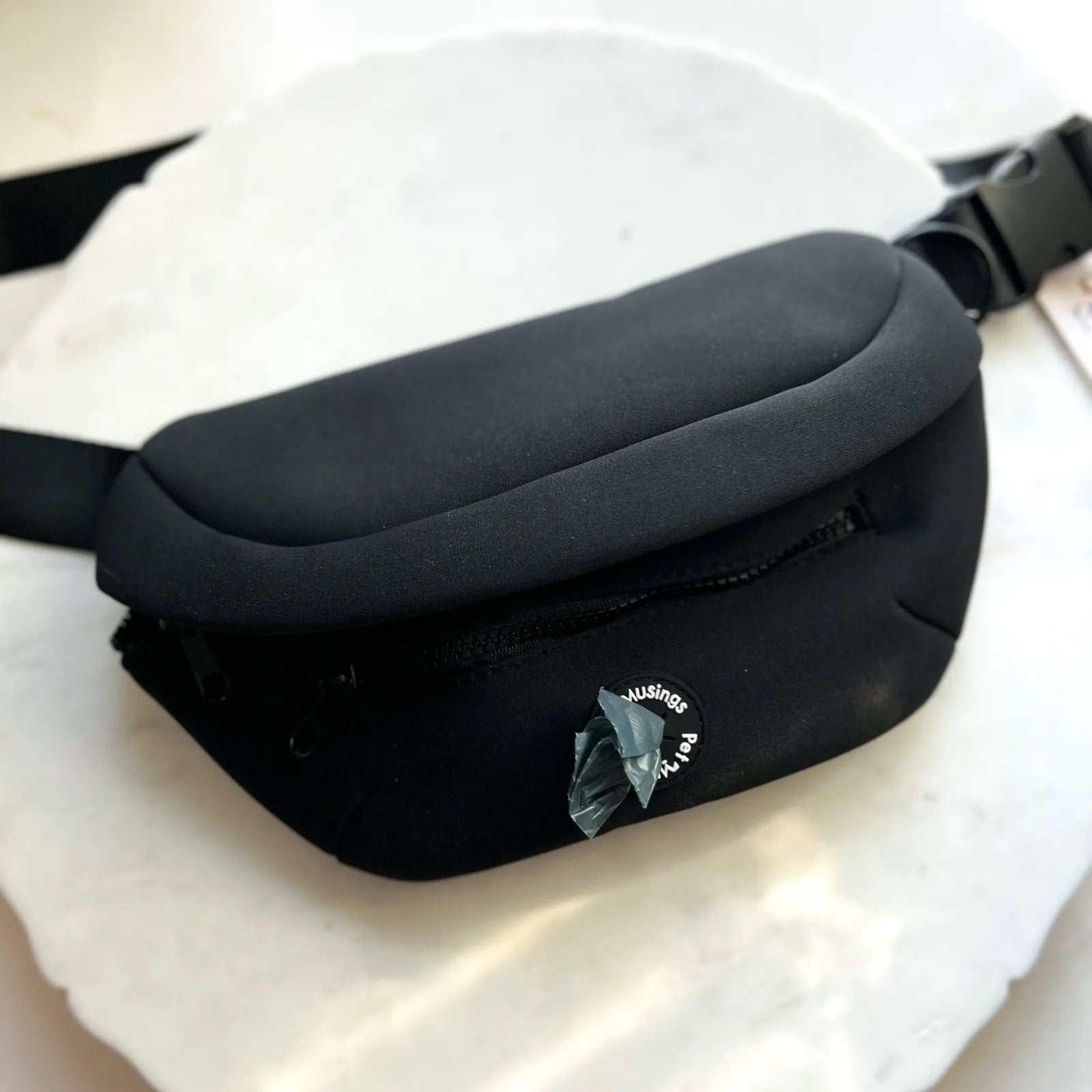 Black fanny pack by Pet Musings, featuring a zippered pocket and an adjustable strap, resting on a marble surface.
