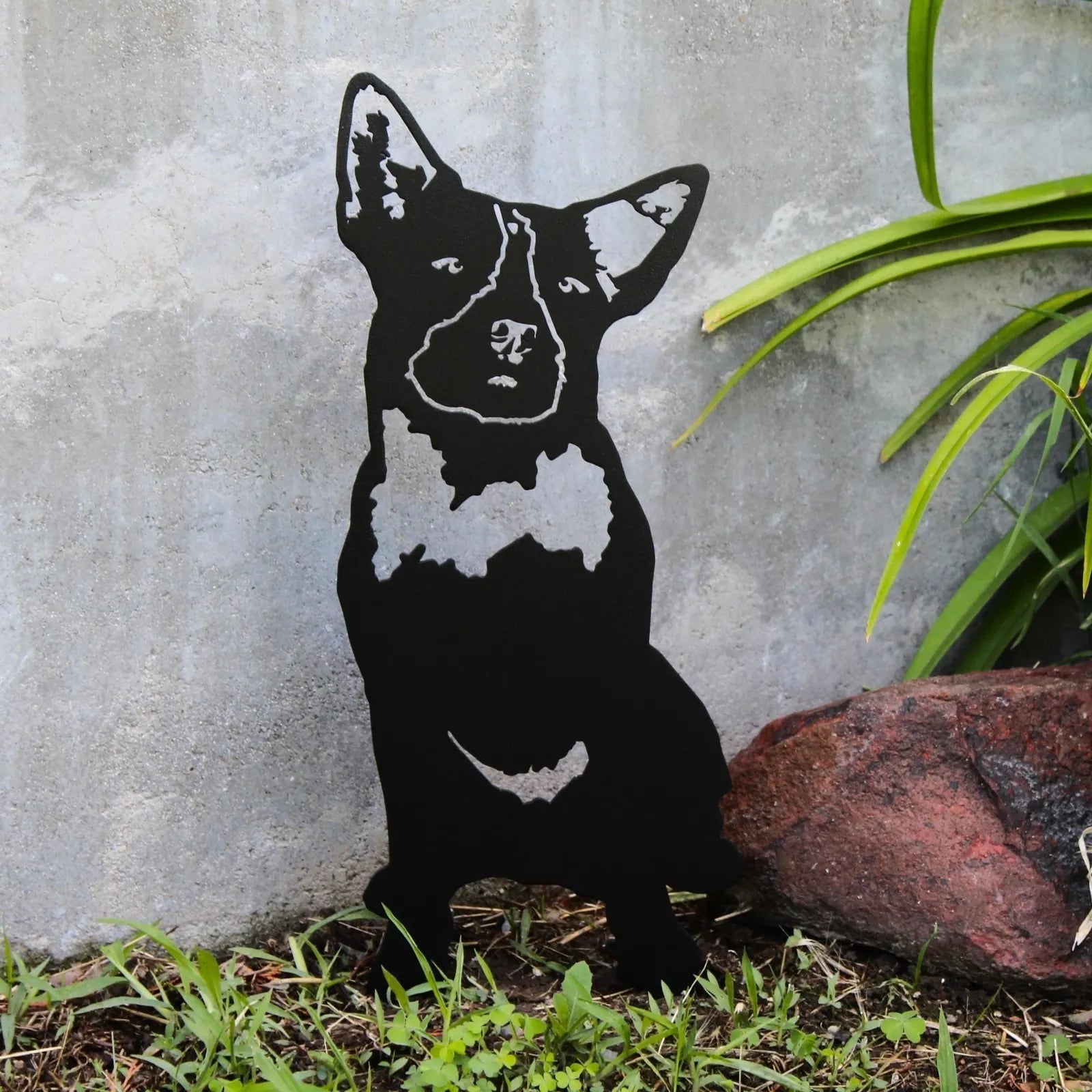 Black metal silhouette of a Blue Heeler dog against a stone wall, surrounded by green grass and garden rocks.