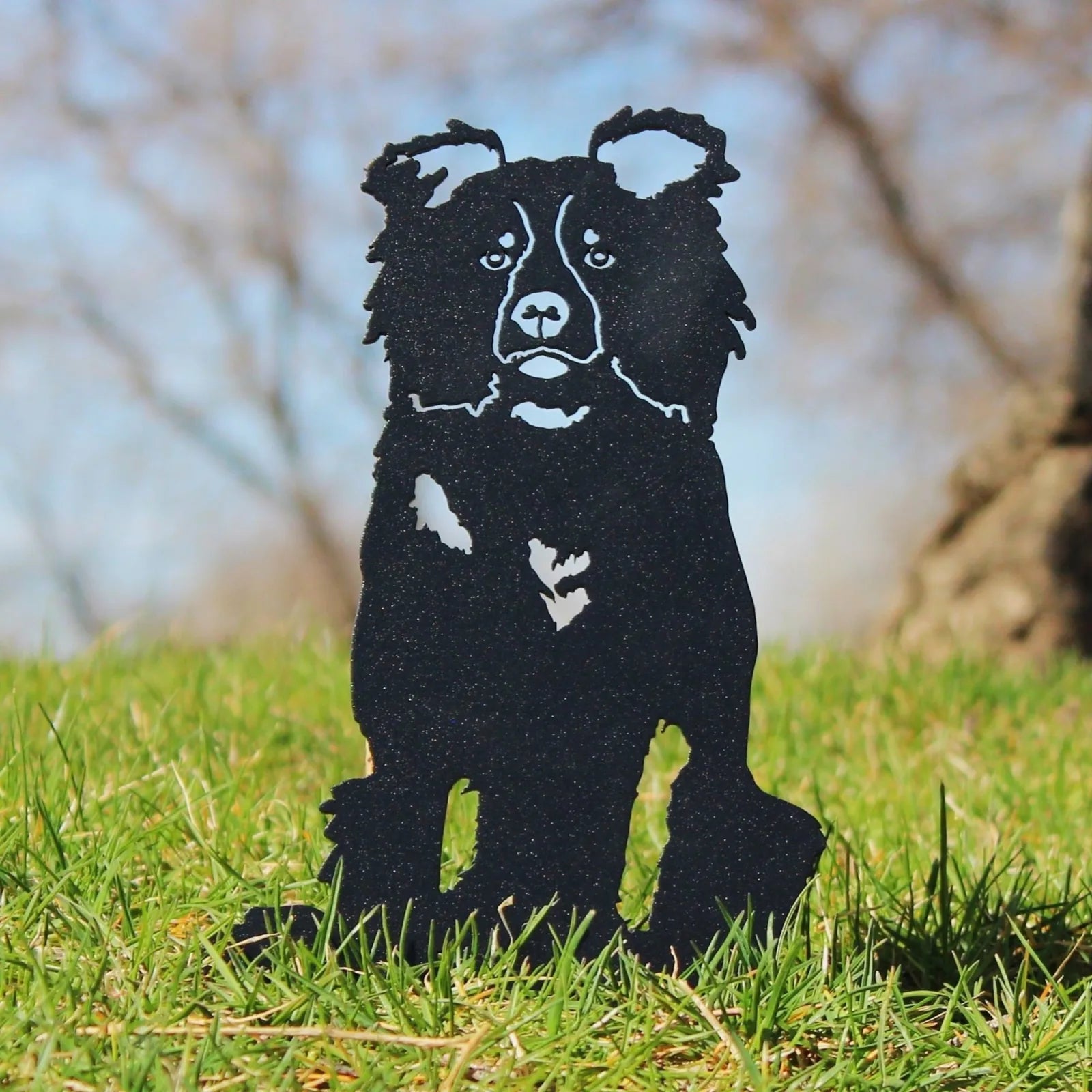 Black metal silhouette of a Border Collie sitting on grass, showcasing detailed features against a natural background.