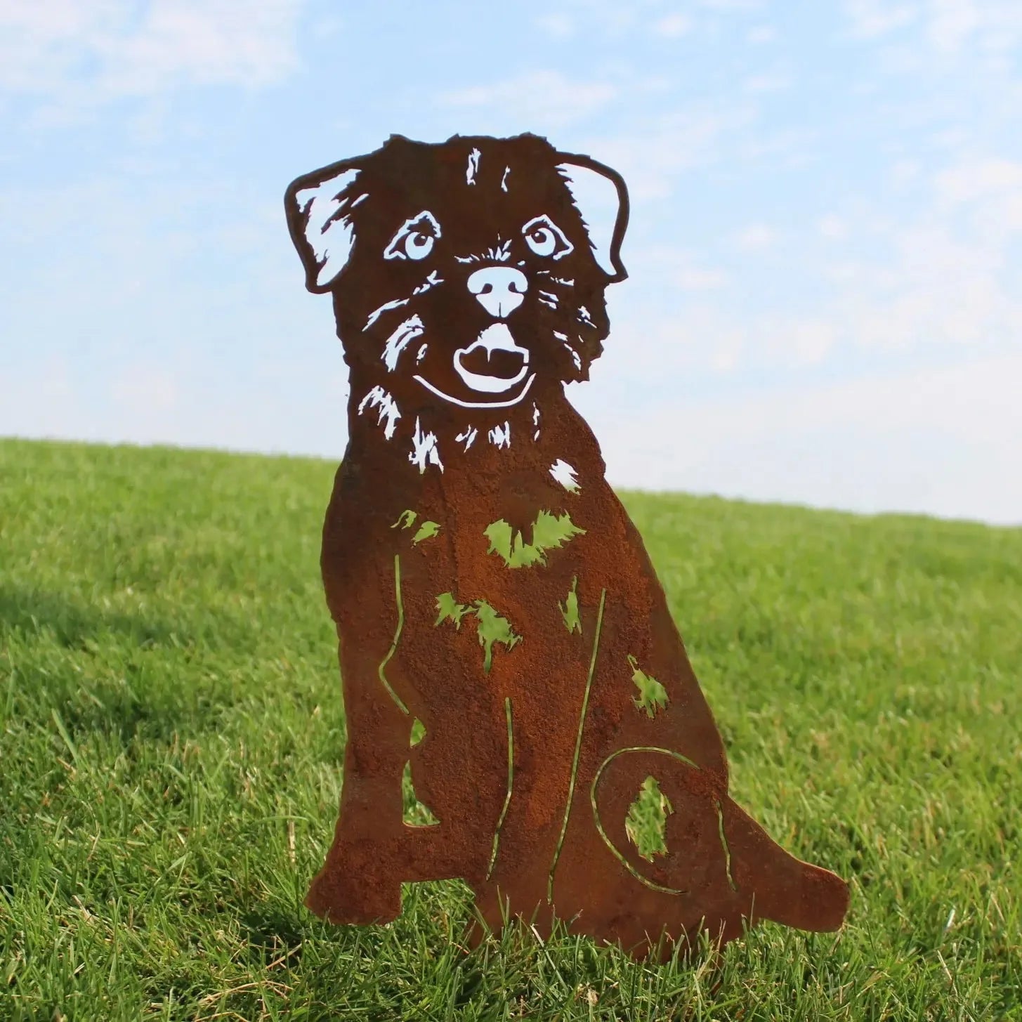 Border Terrier metal silhouette art in rust finish, set against a grassy background under a blue sky.
